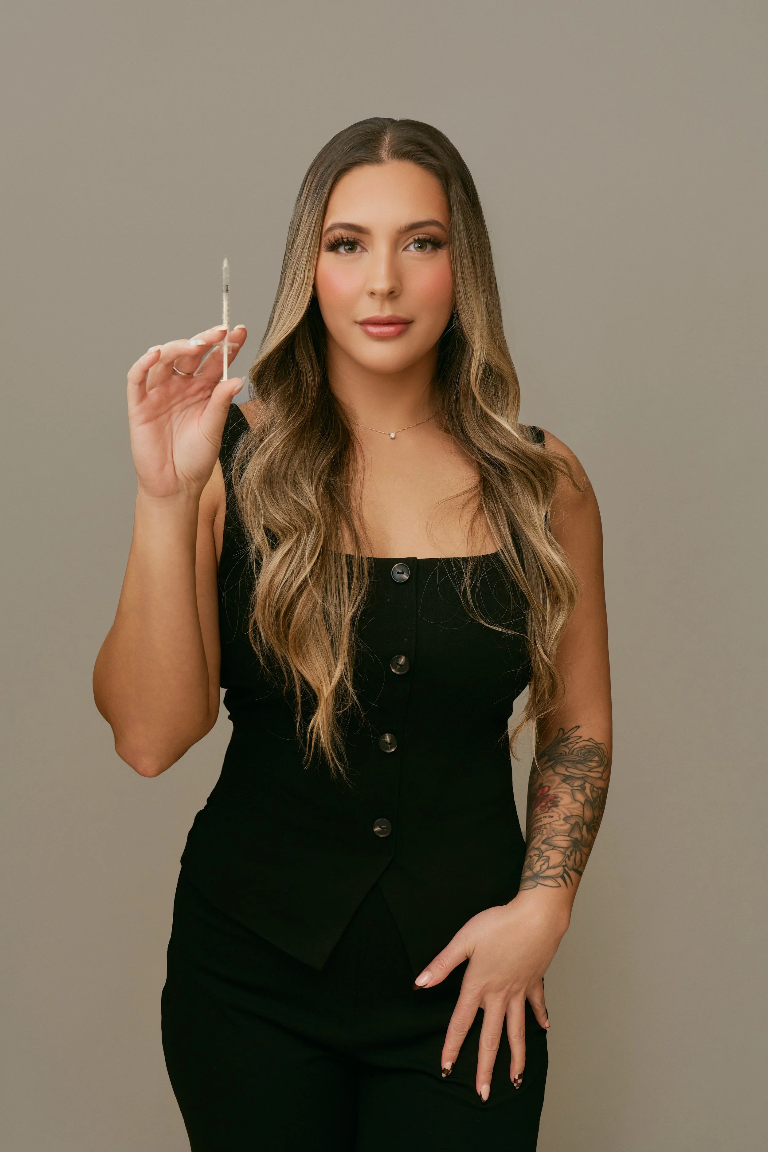 A woman with long wavy hair holding a syringe, dressed in a black button-up dress, standing against a plain gray background.