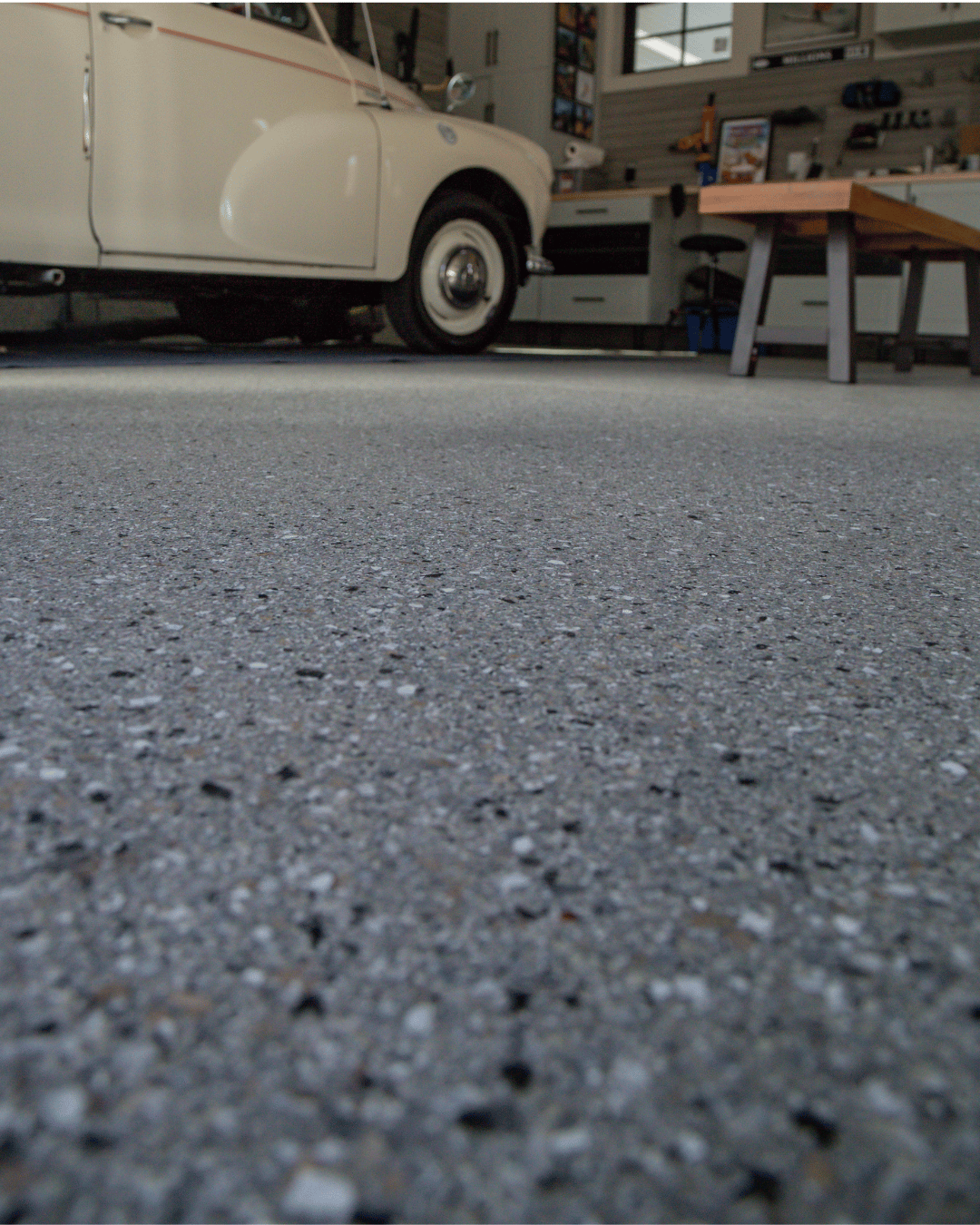 An indoor garage with a vintage cream-colored car parked on a speckled gray concrete floor, with a workbench and tools on the wall in the background.