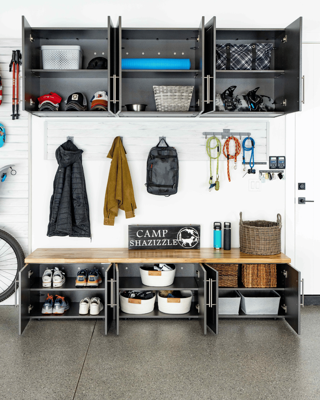 Garage storage area with hooks for jackets, a bench with shoe storage, and upper cabinets containing hats, a yoga mat, and gear for outdoor activities.