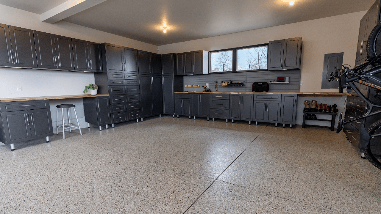 A spacious, organized garage with dark gray cabinets, a wooden workbench, and storage shelves. There is a stool, a potted plant, and various tools. A bicycle hangs on the right side, and windows provide natural light.