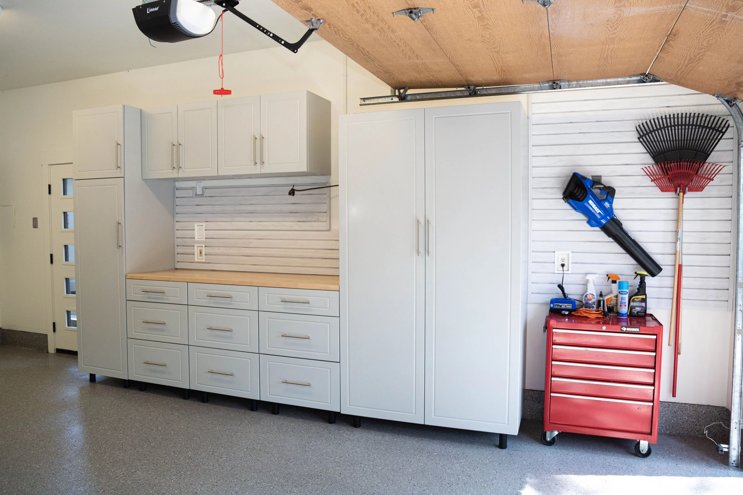 A garage or workshop area with white cabinets, a pegboard, and various tools and cleaning supplies. There is a red tool chest on wheels, a leaf rake, and a handheld vacuum cleaner.