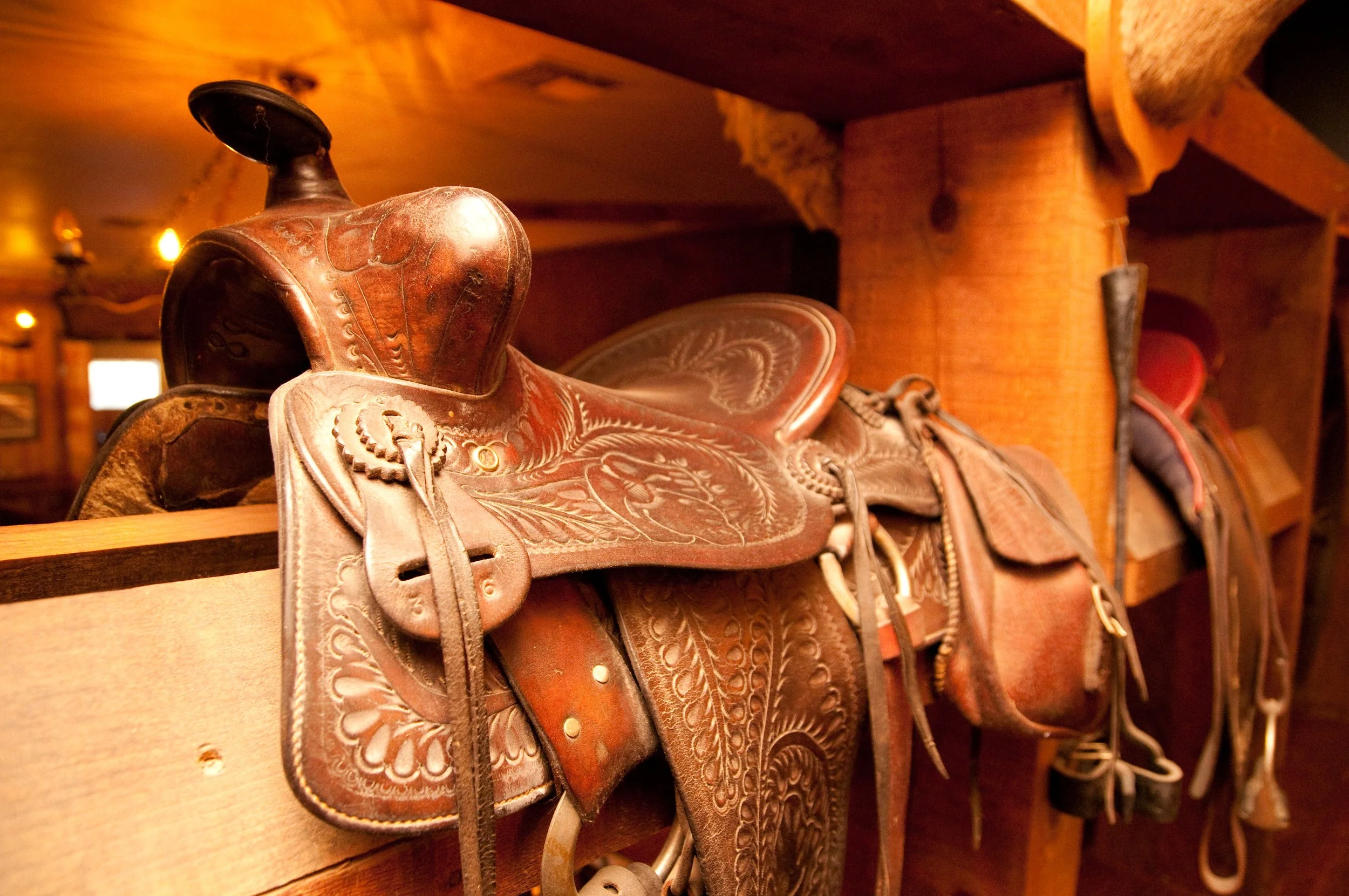 A leather western saddle with intricate tooling resting on a wooden shelf in a rustic setting.
