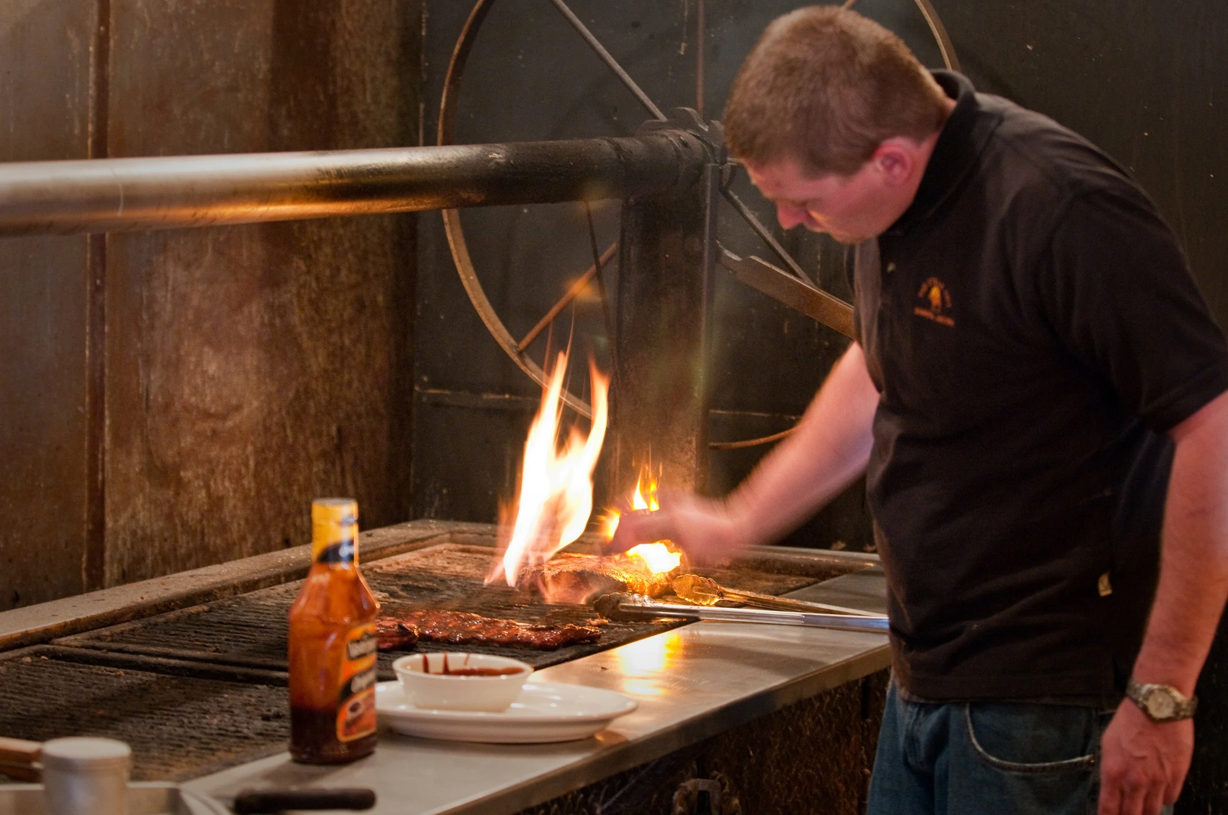 A man cooking meat on a grill with flames, with a bottle of hot sauce and a bowl of sauce on the counter nearby.