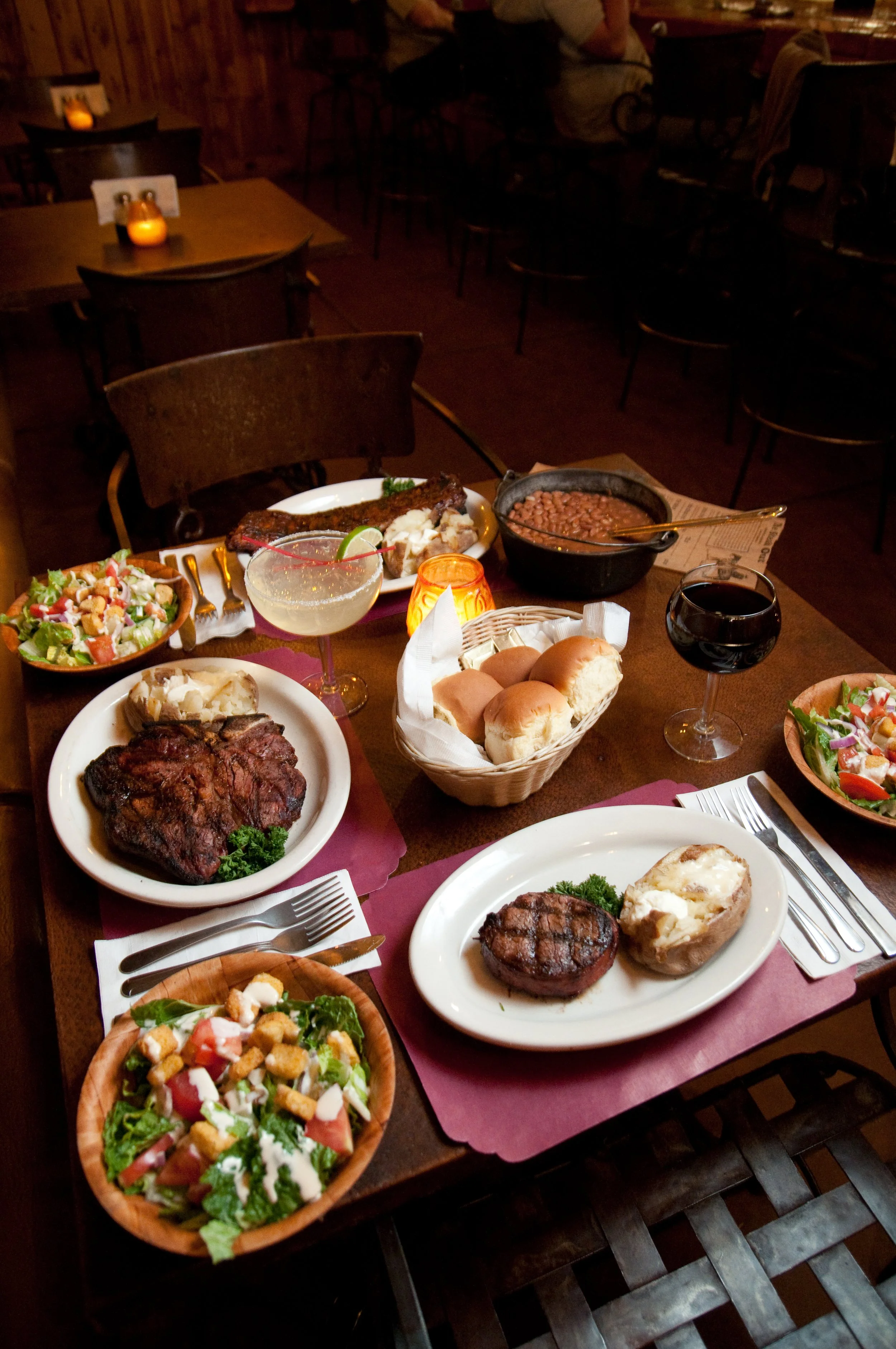 Table filled with various dishes including salad, grilled meat, beans, rolls, and two glasses of wine, in a dimly lit restaurant setting.