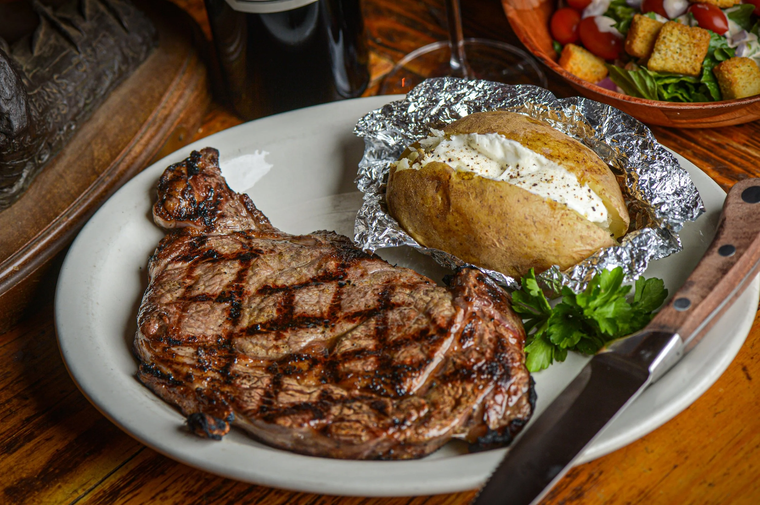 Cooked steak with grill marks on a white plate, topped with a baked potato wrapped in foil and topped with sour cream and black pepper, served with a side salad of cherry tomatoes, croutons, and lettuce in a bowl, all on a wooden table.