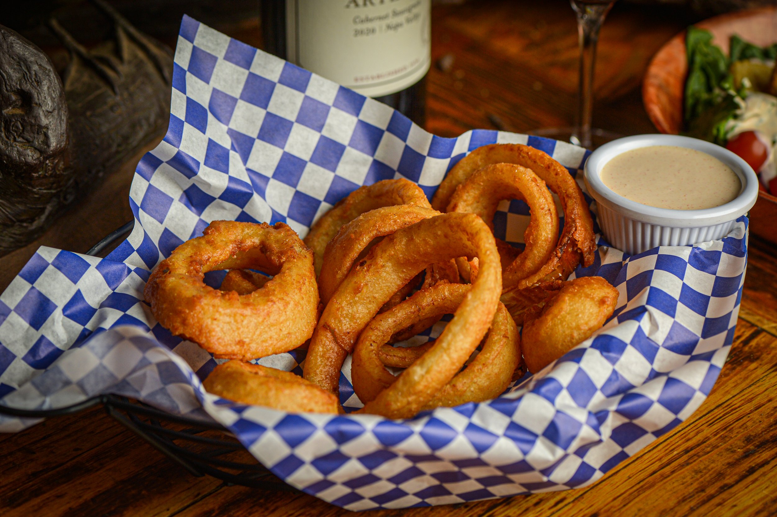 Basket of fried onion rings with a side of creamy dipping sauce, on a checkered paper lining, with a salad, a glass of wine, and a bottle in the background.