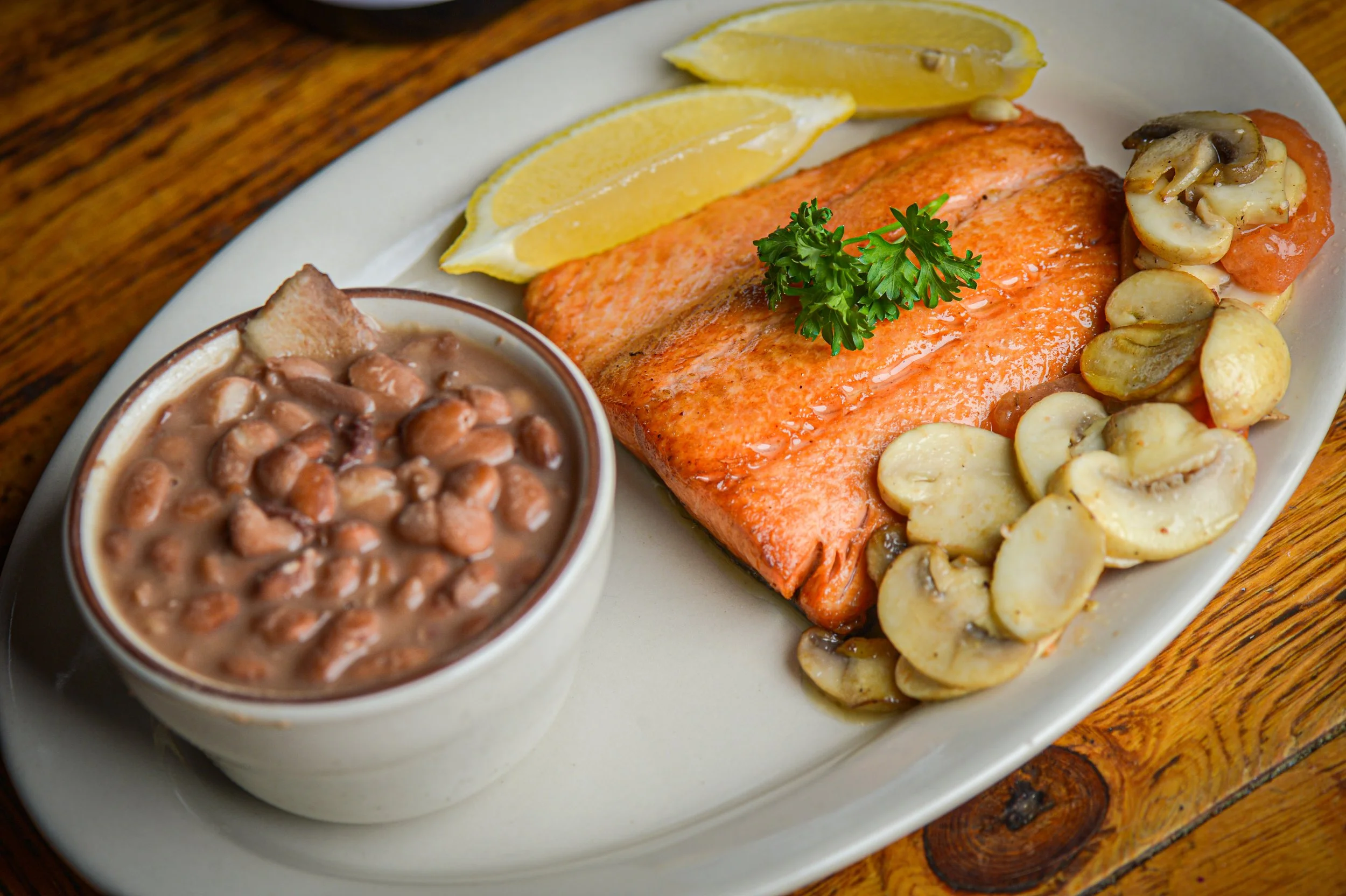 Plate with cooked salmon, lemon wedges, mushroom and potato side dishes, garnished with parsley, on a wooden surface.