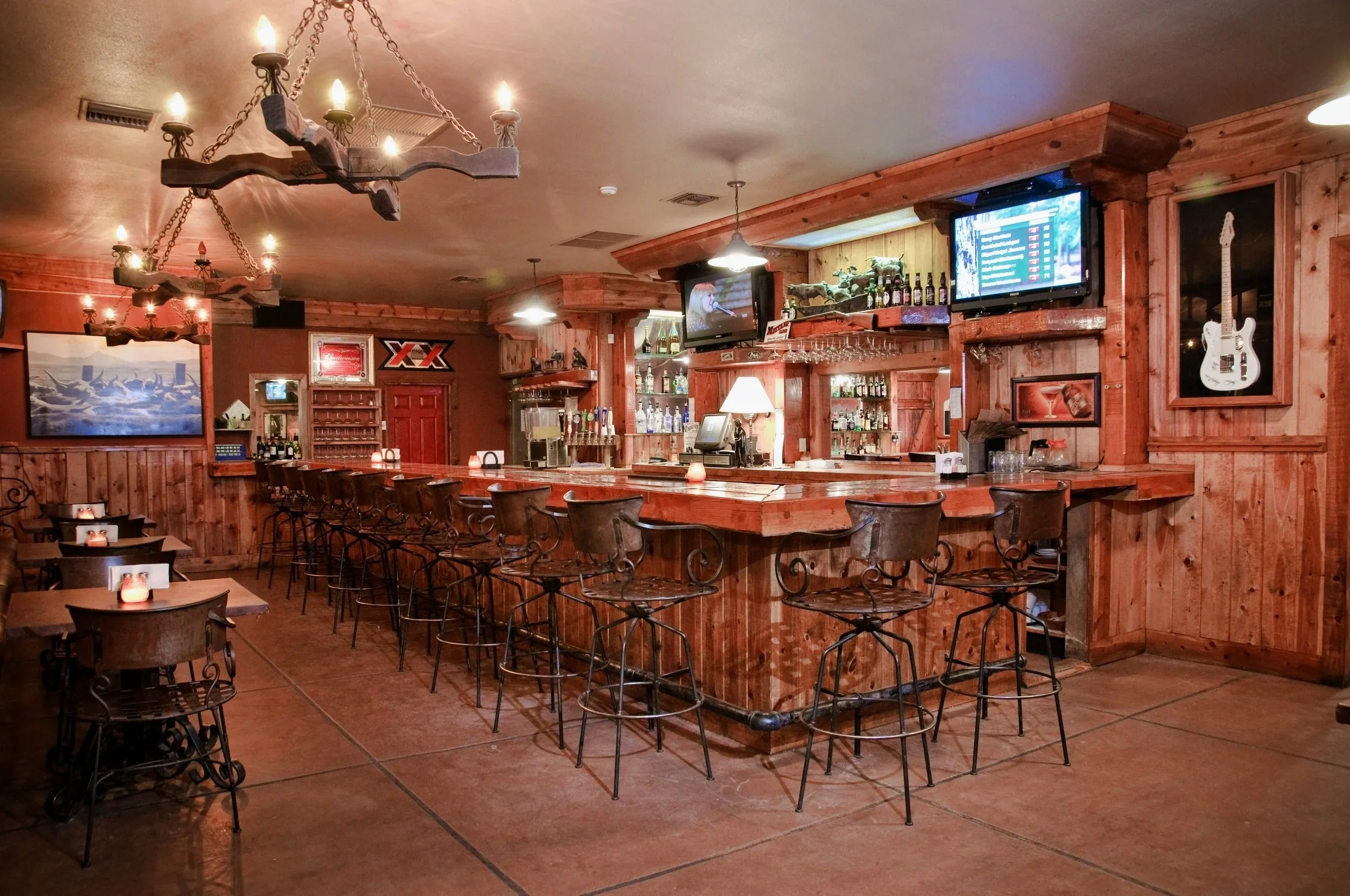 Interior of a bar with a wooden counter, bar stools, and mounted TV screens showing a person singing. The bar is decorated with guitars on the wall, pictures, and decorative items, with warm lighting and chandeliers.