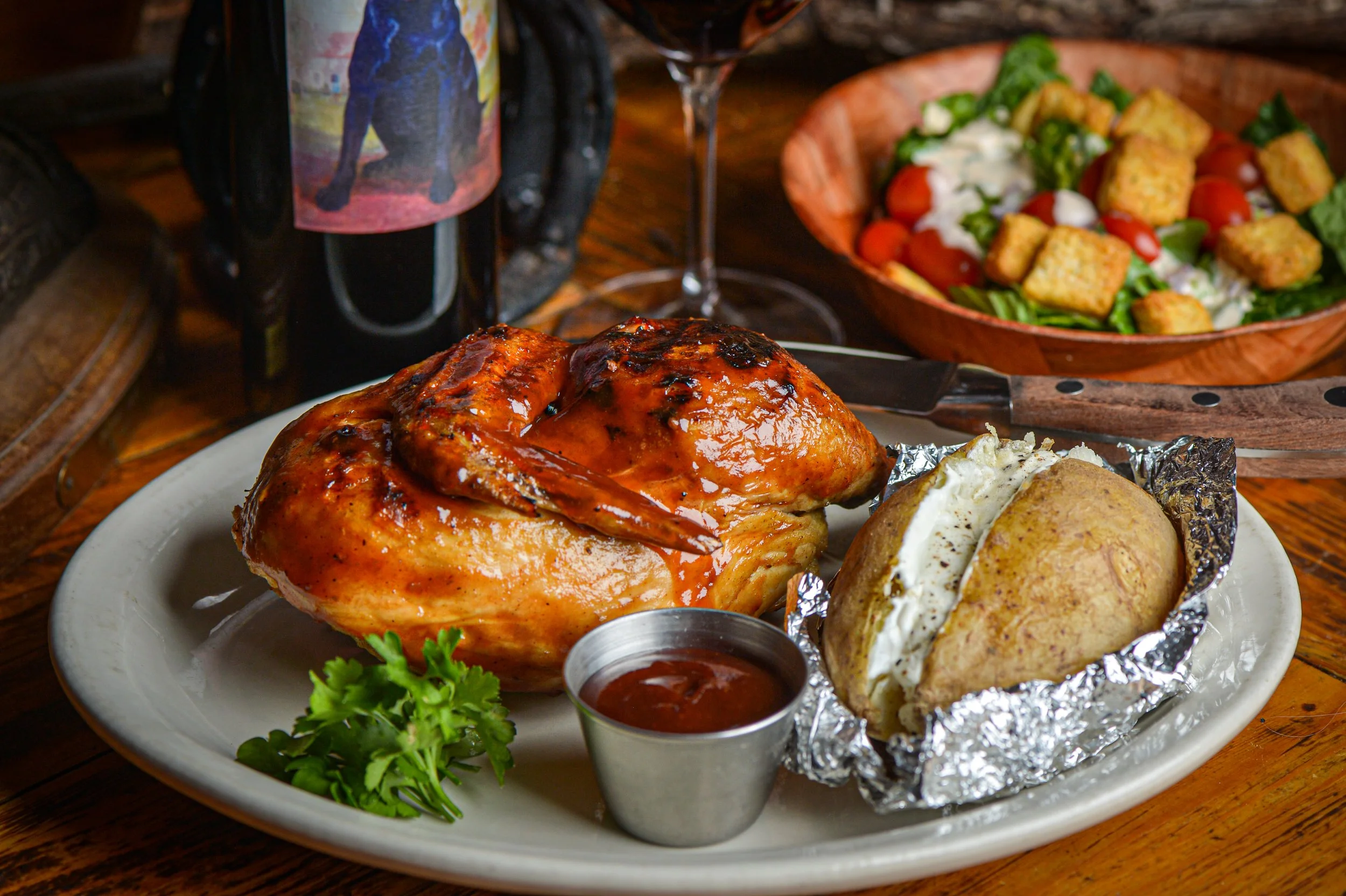 Baked chicken with barbecue sauce served with baked potato stuffed with sour cream and chives, alongside a small cup of barbecue sauce, a leafy garnish, and a salad with cherry tomatoes, cucumber, and croutons in a wooden bowl.