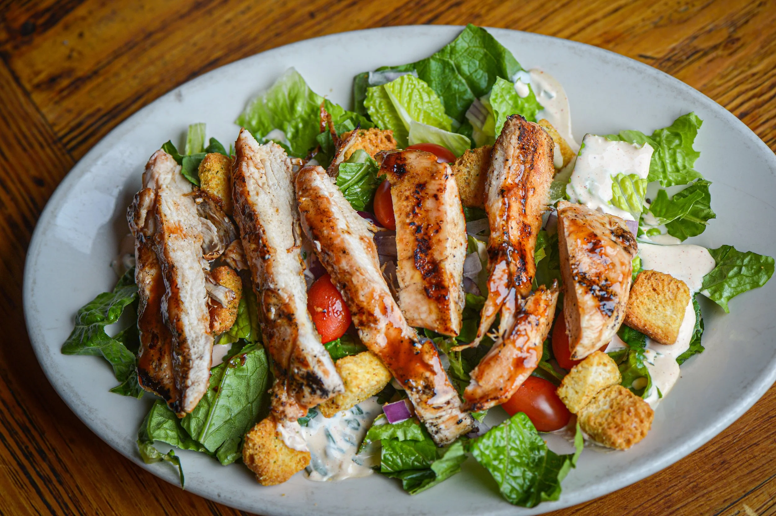 Grilled chicken salad with lettuce, cherry tomatoes, croutons, shredded cheese, and dressing on a white plate