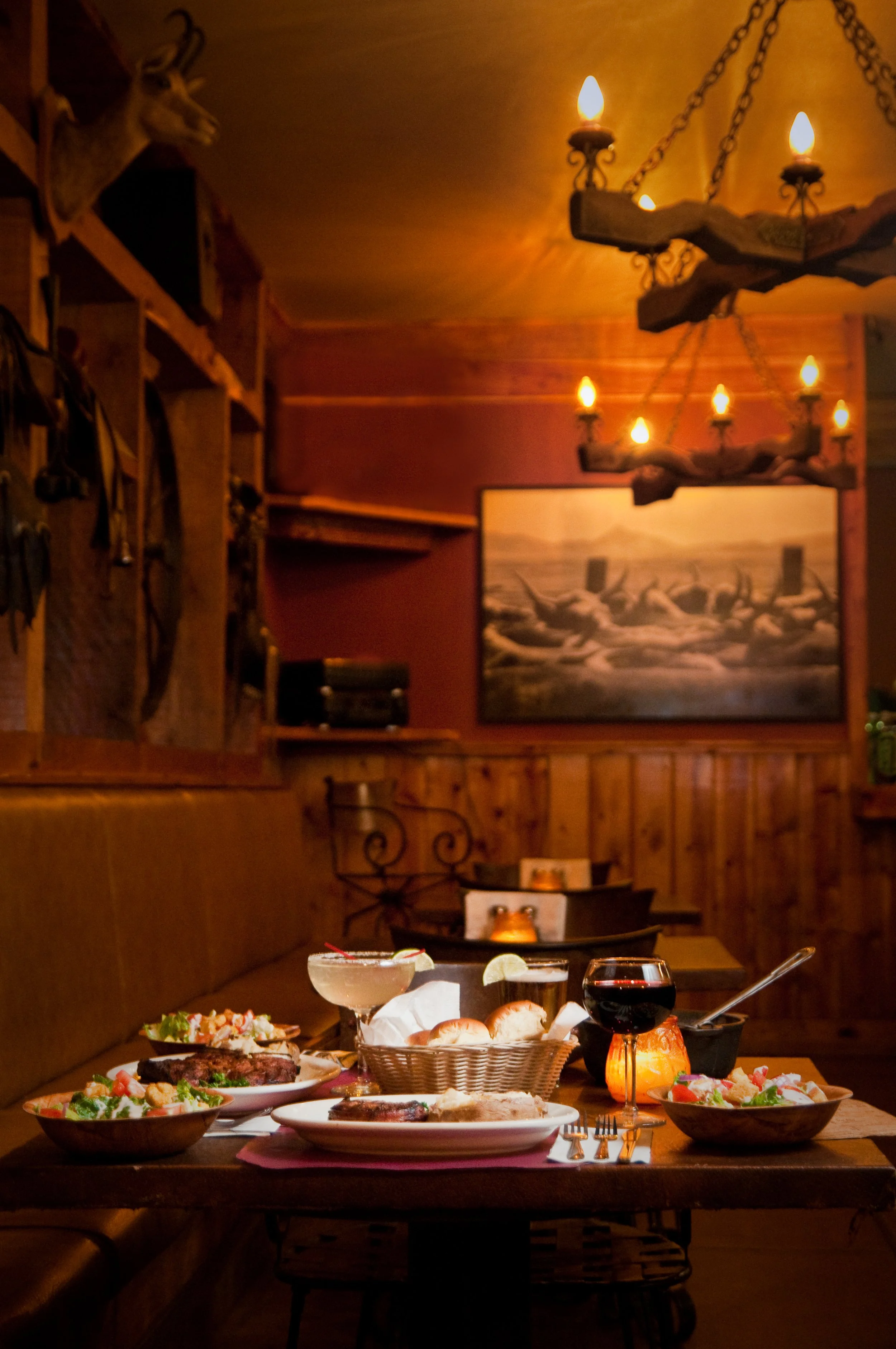 A cozy restaurant setting with a table full of various dishes, glasses of wine, and a candle in a small lantern. The background features warm lighting, a large painting, and rustic decor including a mounted deer head and a wooden chandelier.