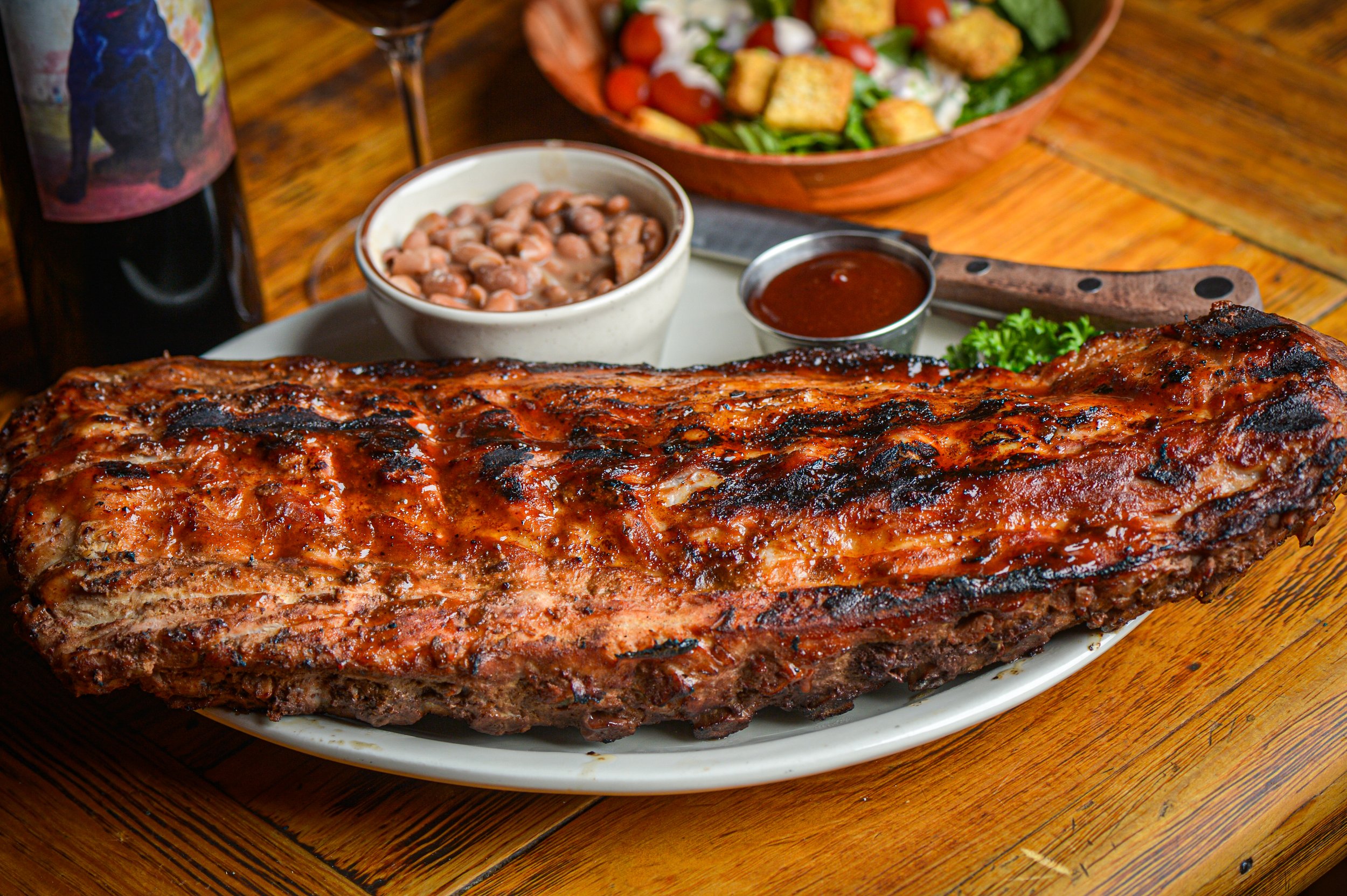 Barbecue pork ribs with a glaze, served on a white plate with a side of baked beans and barbecue sauce, accompanied by a salad in a wooden bowl and a bottle of beverage in the background.