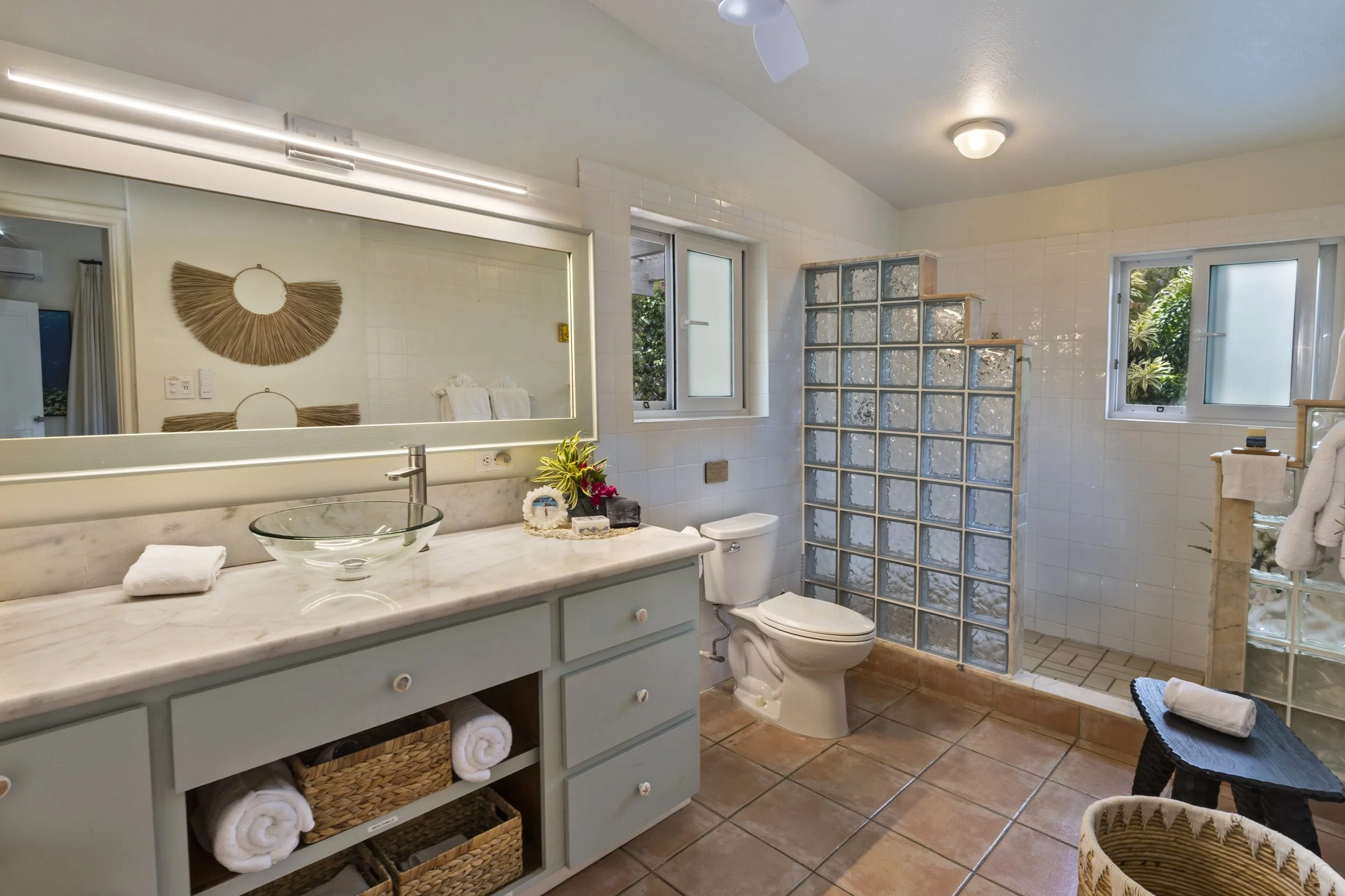Mandavilla St. John USVI vacation rental A spacious bathroom with a large mirror, a marble countertop, windows. There is a glass block wall separating the shower area, a white toilet, and a wicker laundry basket with a towel on it.