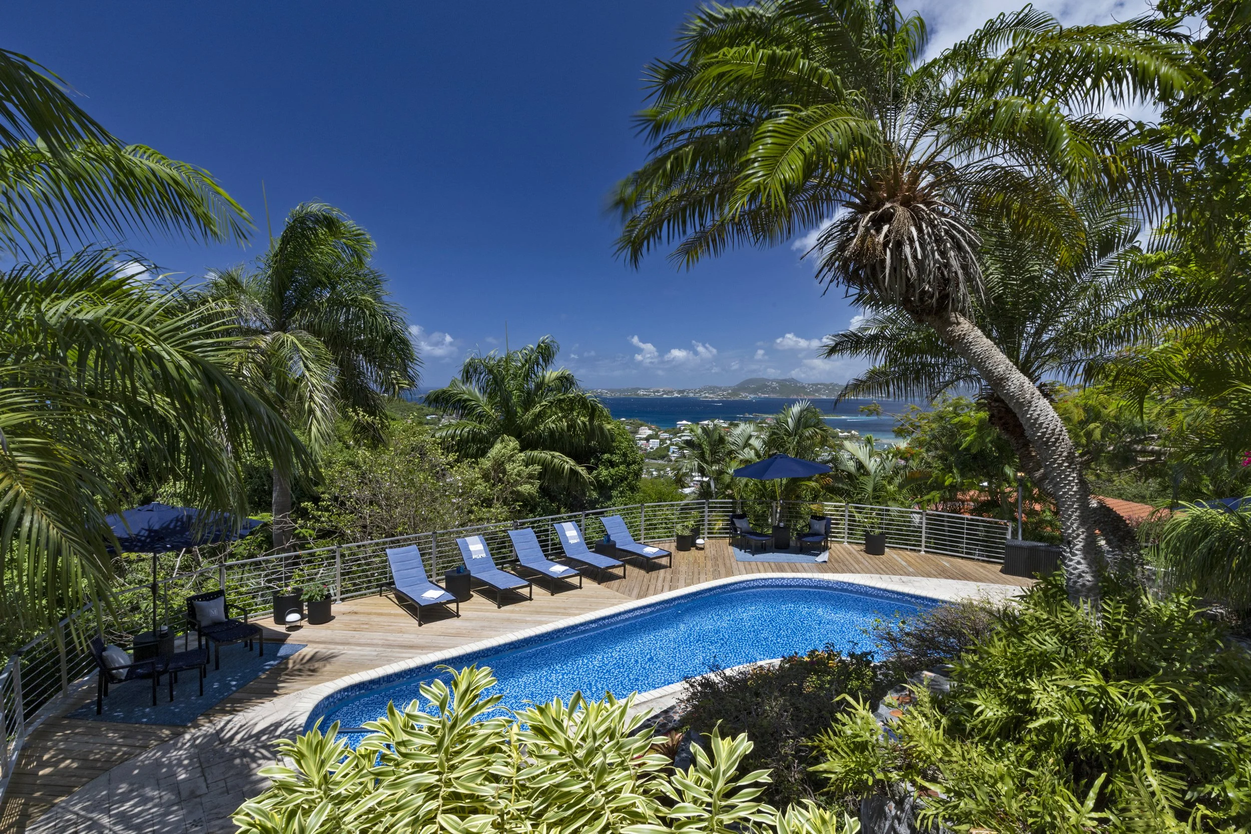 Mandavilla St. John USVI vacation rental A rooftop pool area surrounded by tropical palm trees with a view of the ocean and distant islands in the background, under a blue sky.