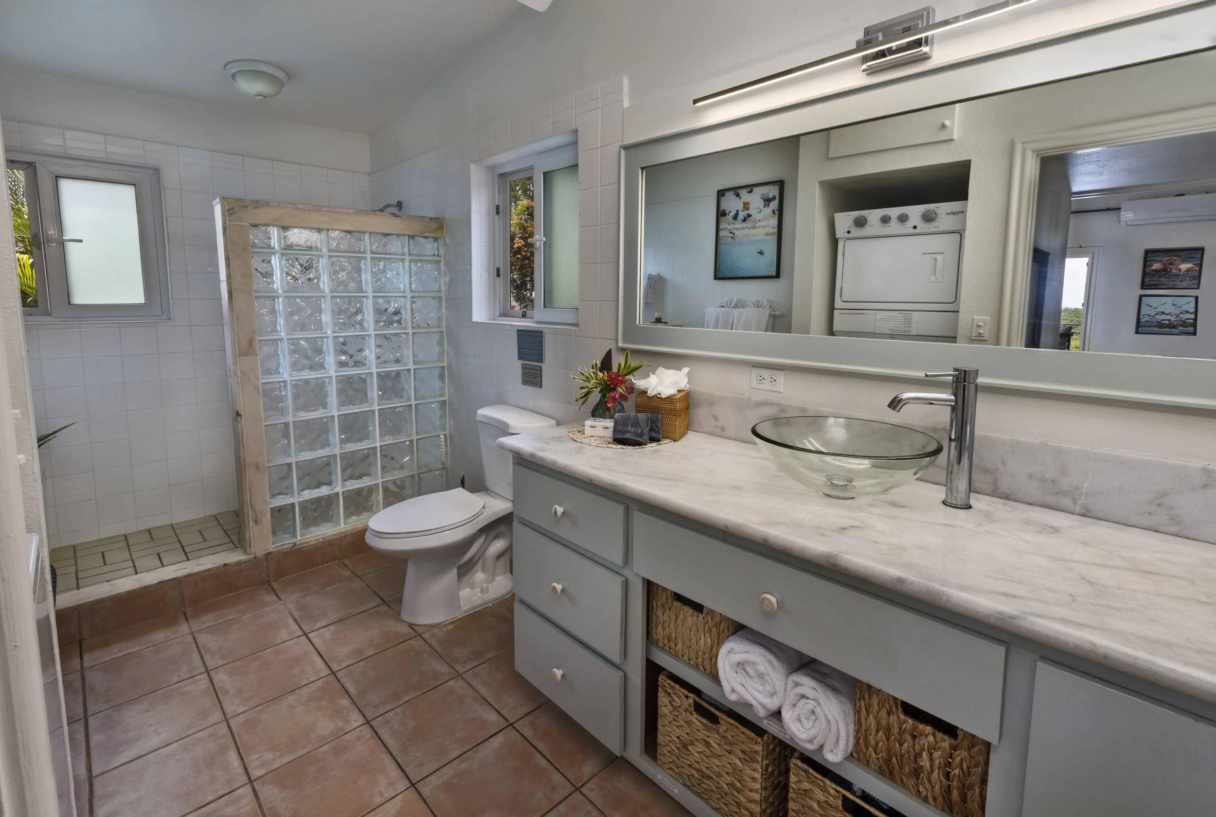 Mandavilla St. John USVI vacation rental Bathroom with a glass bowl sink on a marble countertop, a large mirror, laundry machine, shower area with glass block wall, window, and various towels and decorative items.