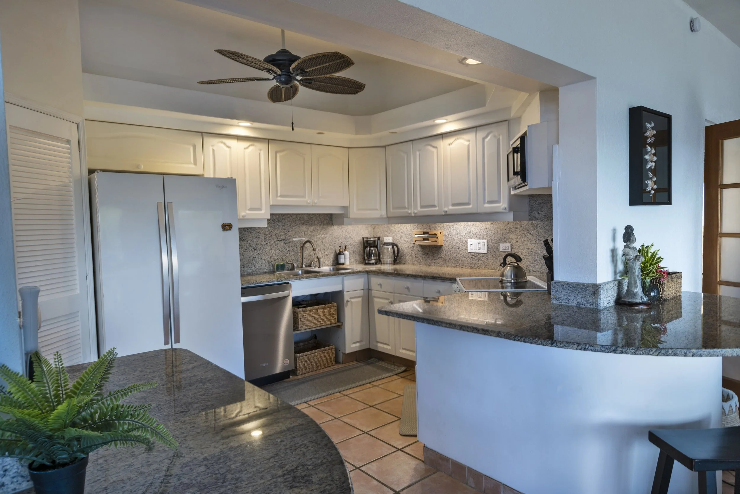 Mandavilla St. John USVI vacation rental A kitchen with white cabinets, granite countertops, a stainless steel fridge, coffee maker, and kitchenware. There is a ceiling fan and decorative plant on the counter.