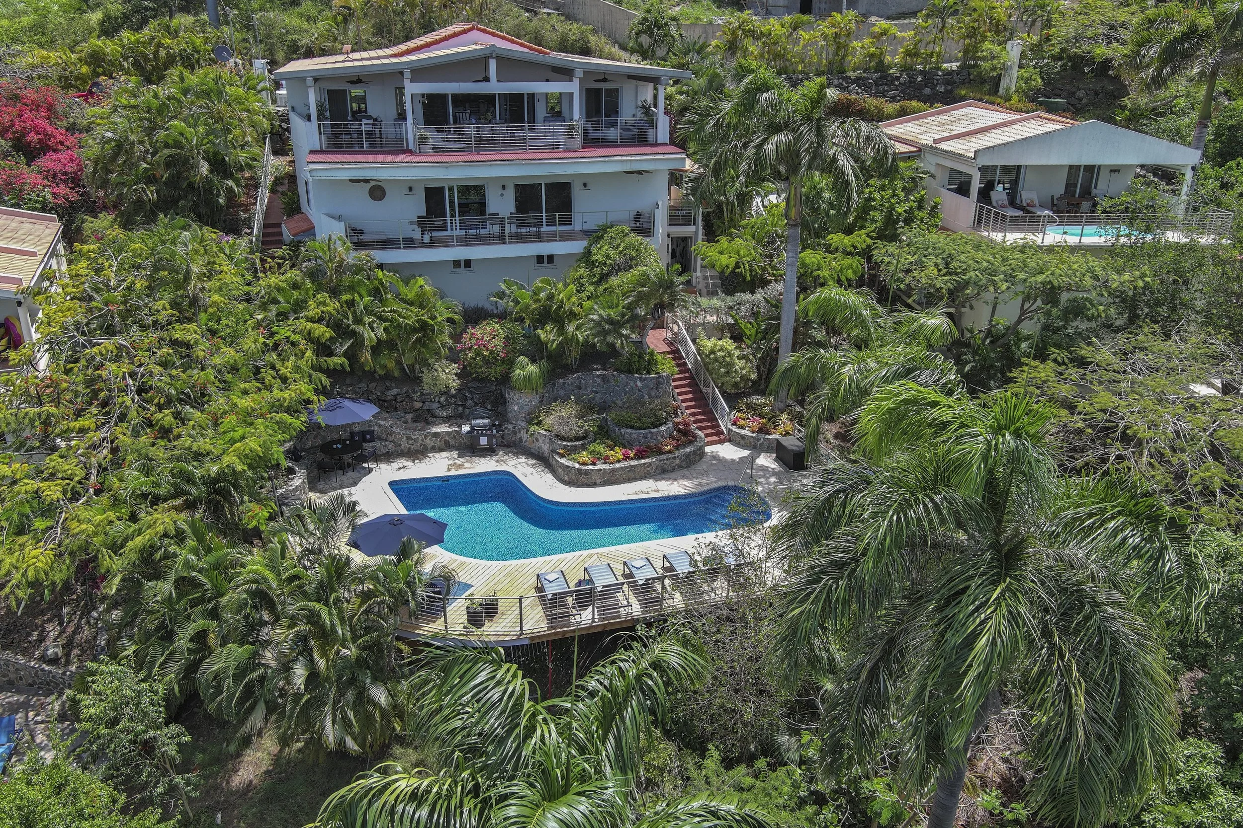 Mandavilla St. John USVI vacation rental Aerial view of a tropical residential area featuring a large white house with multiple decks, a backyard with a shaped swimming pool, surrounded by lush green palm trees and vibrant flowering plants.