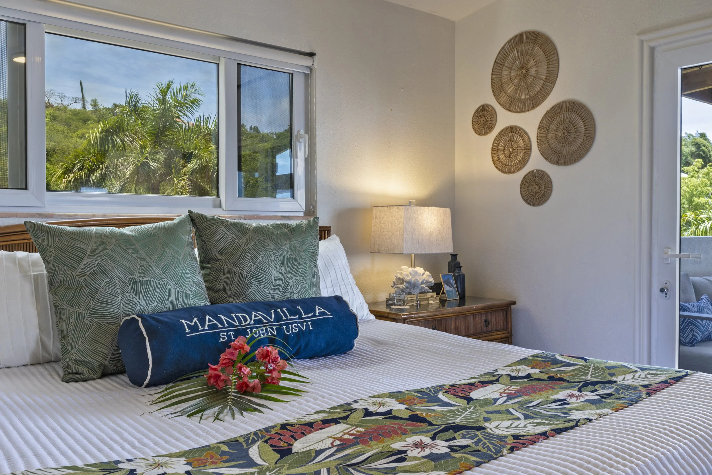 Mandavilla St. John USVI vacation rental cozy bedroom with a bed topped with green leaf-patterned pillows, a white duvet, a floral bed runner, and decorative wall art. Large windows show lush greenery outside.