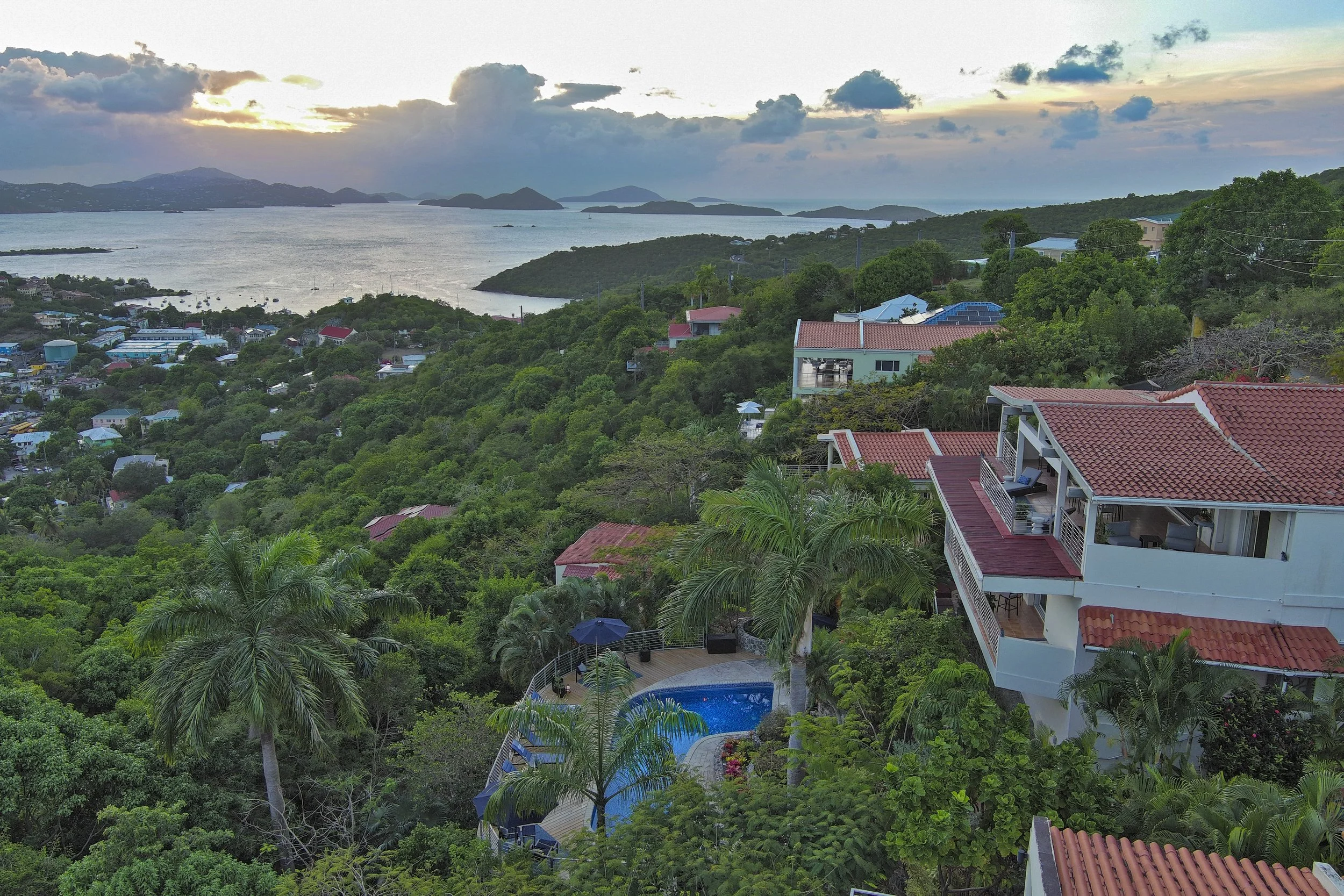 Mandavilla St. John USVI vacation rental A scenic view of a coastal area with a hillside filled with greenery, houses, and pools. The ocean with several islands and boats is in the background, under a partly cloudy sky.