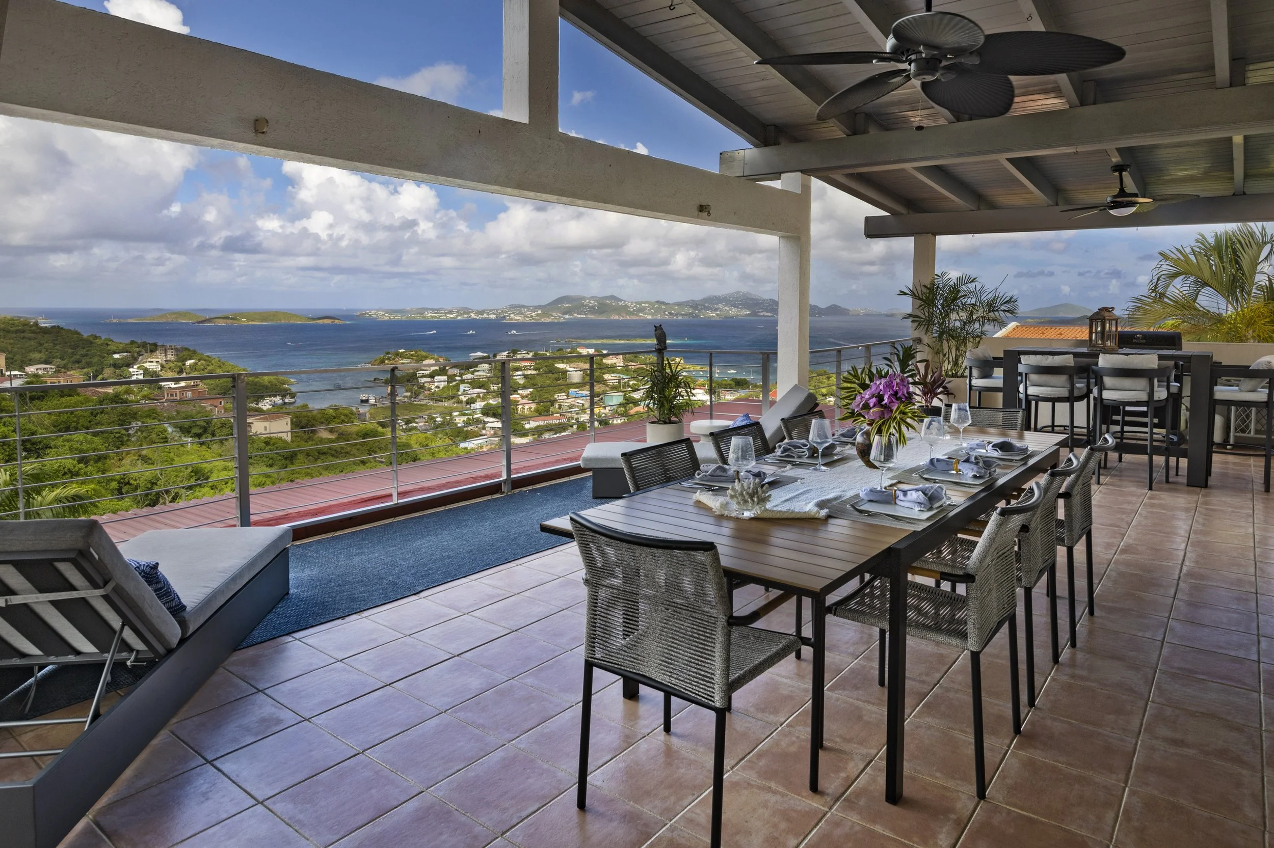 Mandavilla St. John USVI vacation rental An outdoor balcony dining area with a view of a bay, islands, and hills, featuring a table set for a meal with wine glasses, napkins, and a centerpiece of purple flowers, with ceiling fans and potted plants.