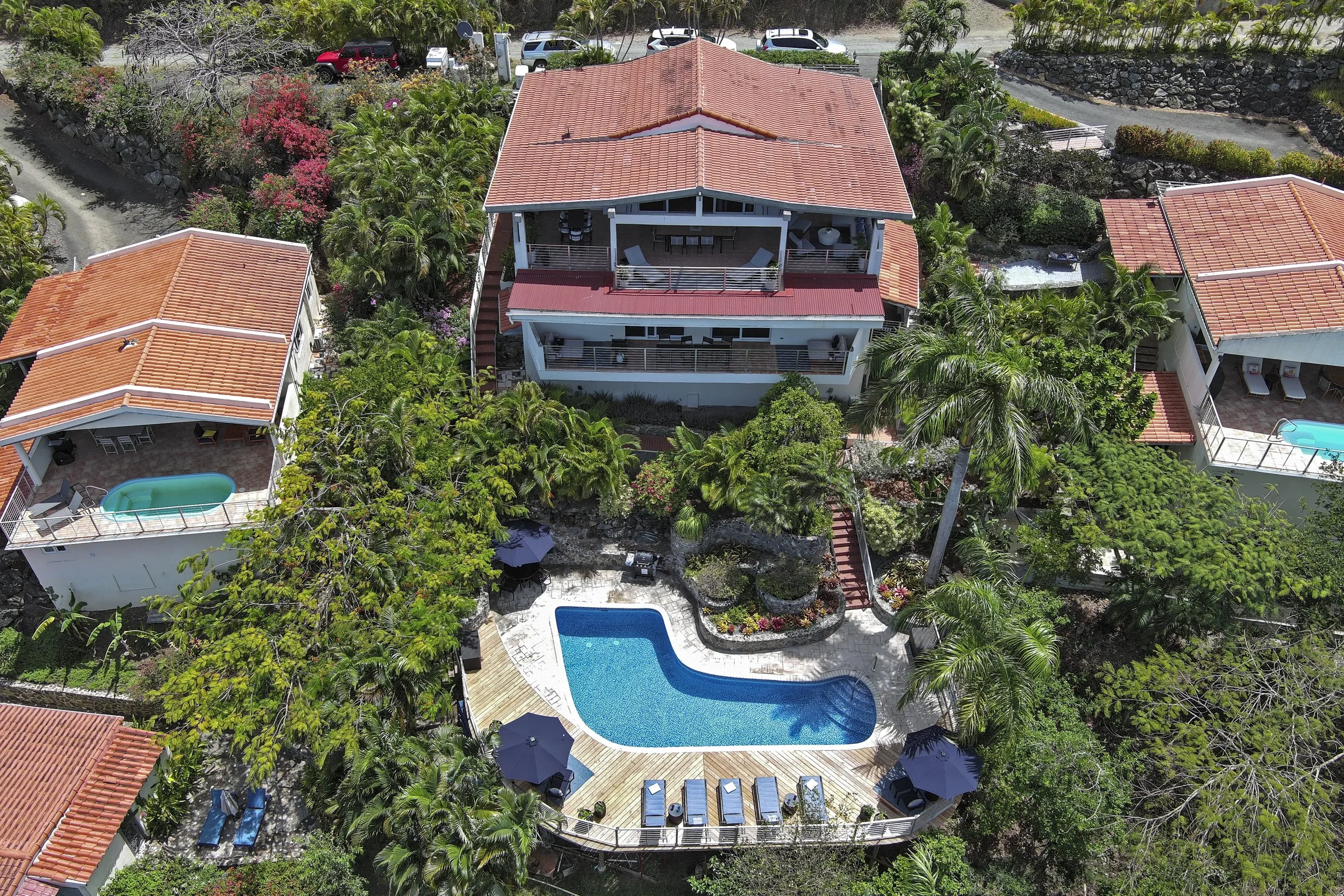Mandavilla St. John USVI vacation rental An aerial view of a large house with red tiled roofs, surrounded by lush green trees and plants, with a swimming pool and poolside lounge chairs in the backyard.