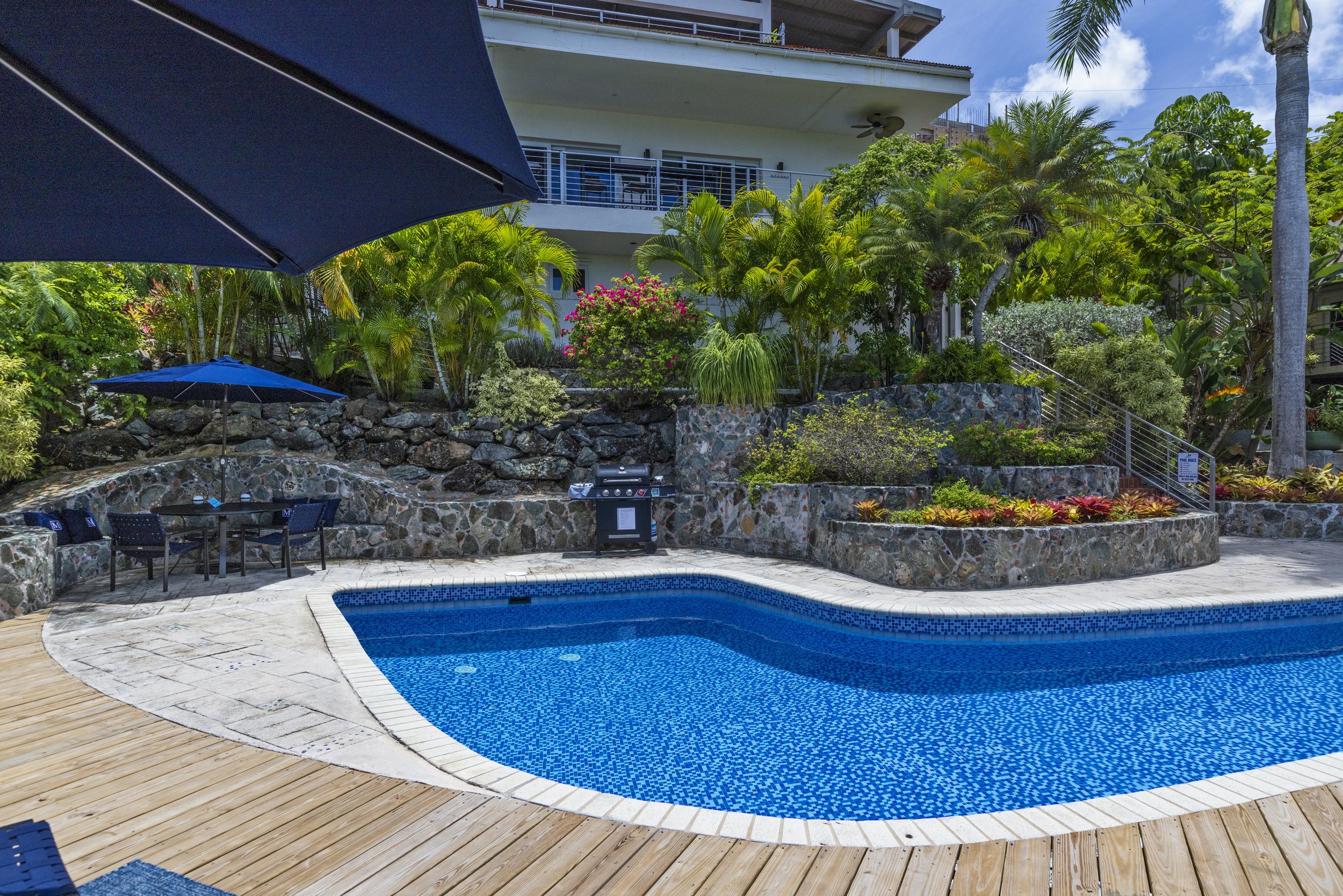 Mandavilla St. John USVI vacation rental An outdoor swimming pool area with a partially visible blue umbrella, a variety of lush green tropical plants, decorative rocks, and a multi-story building in the background. 
