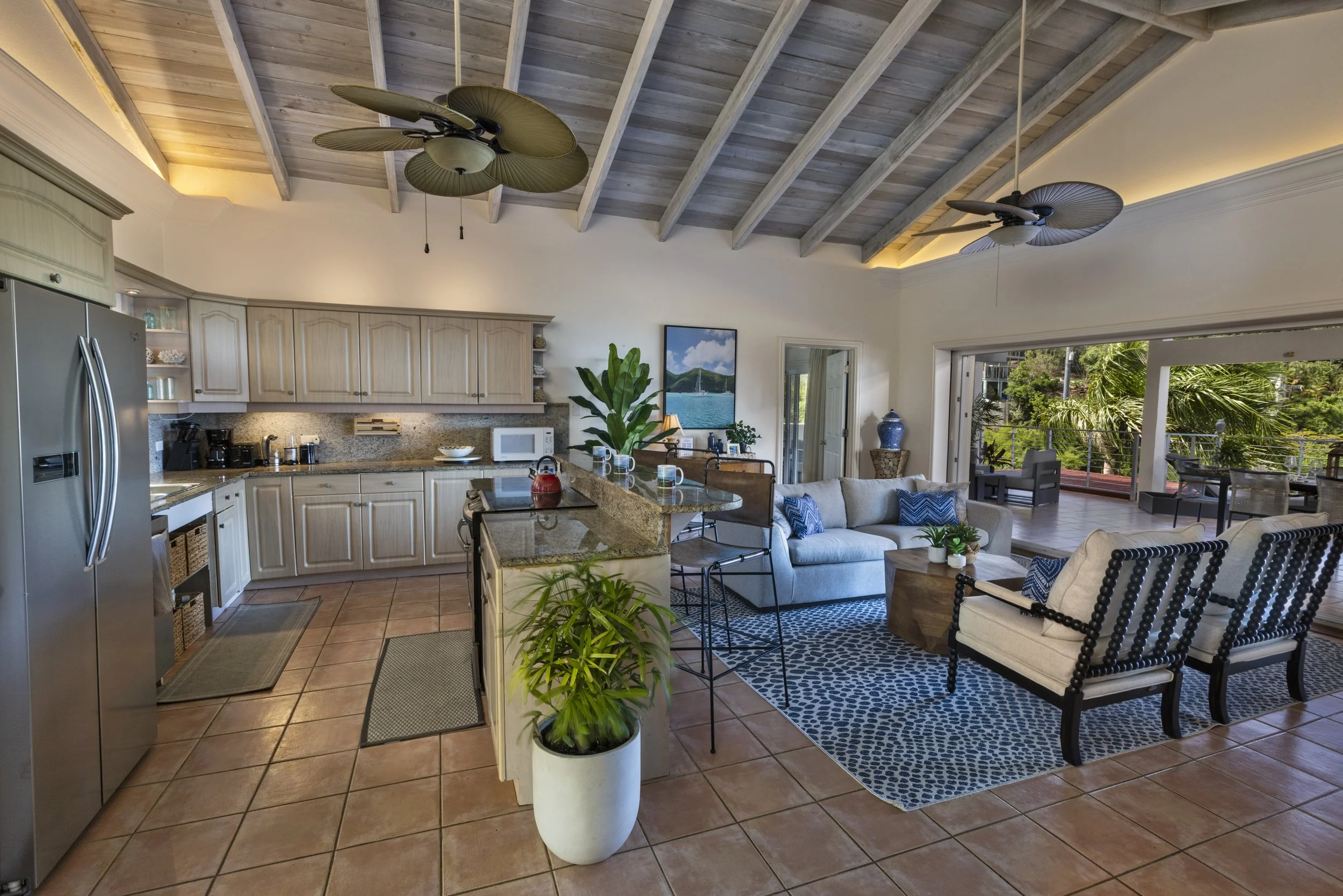 Mandavilla St. John USVI vacation rental A living room and kitchen area with a vaulted wood ceiling, ceiling fans, beige cabinets, a tile floor, and sliding glass doors leading to an outdoor patio with greenery.