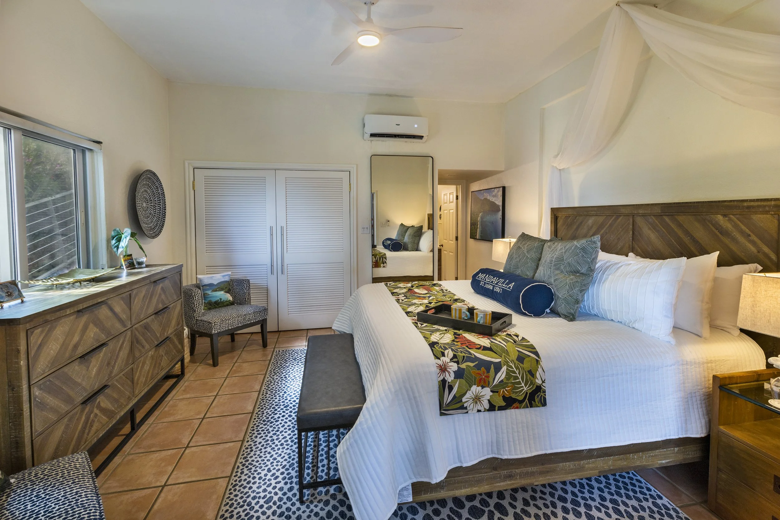 A bedroom with a large bed, decorative pillows, a flower-patterned blanket, a wooden headboard, and a nightstand with a lamp. There's a mirror on the wall, a dresser, a chair, and an open closet door. The ceiling has a fan and an air conditioning uni