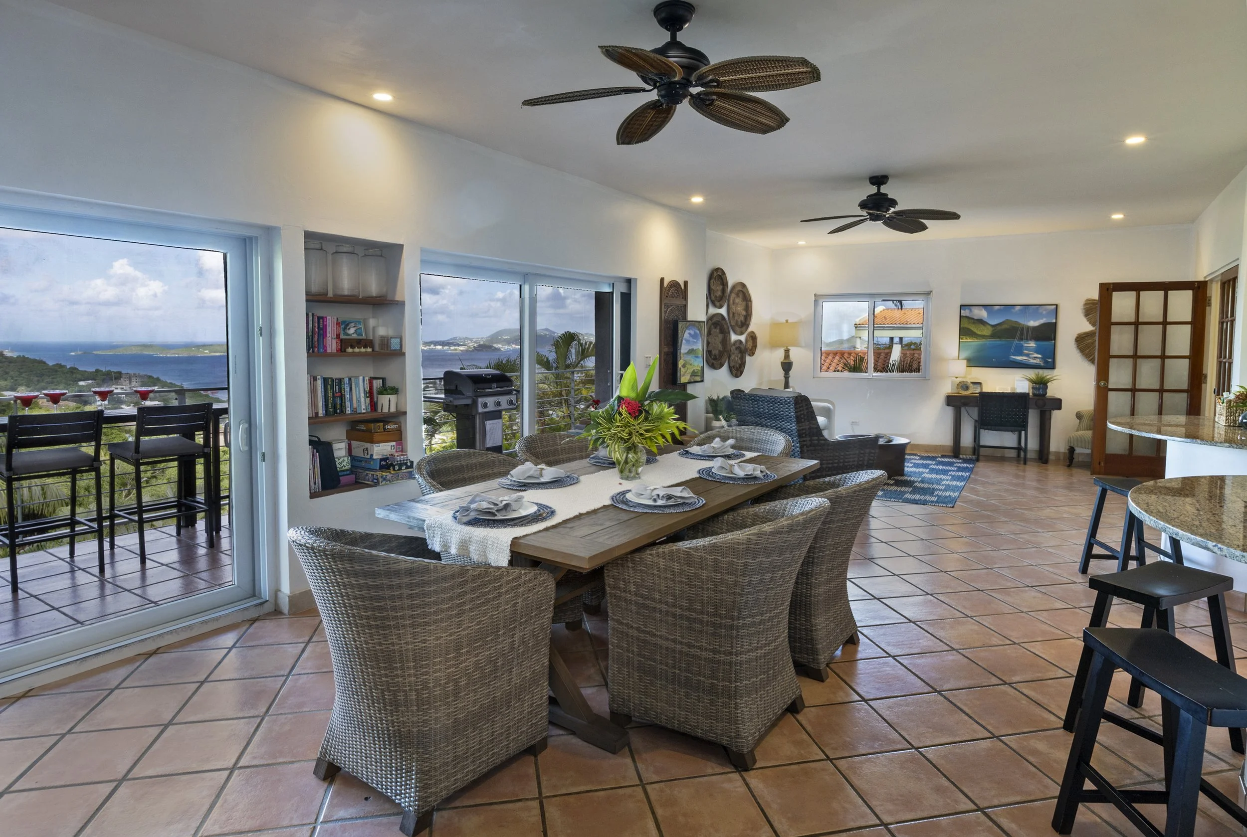 Living room with large windows showing ocean view, dining table with six chairs and a floral centerpiece, bookshelves, wicker chairs, TV, artwork, ceiling fans, clay tile floor, patio with chairs and ocean view