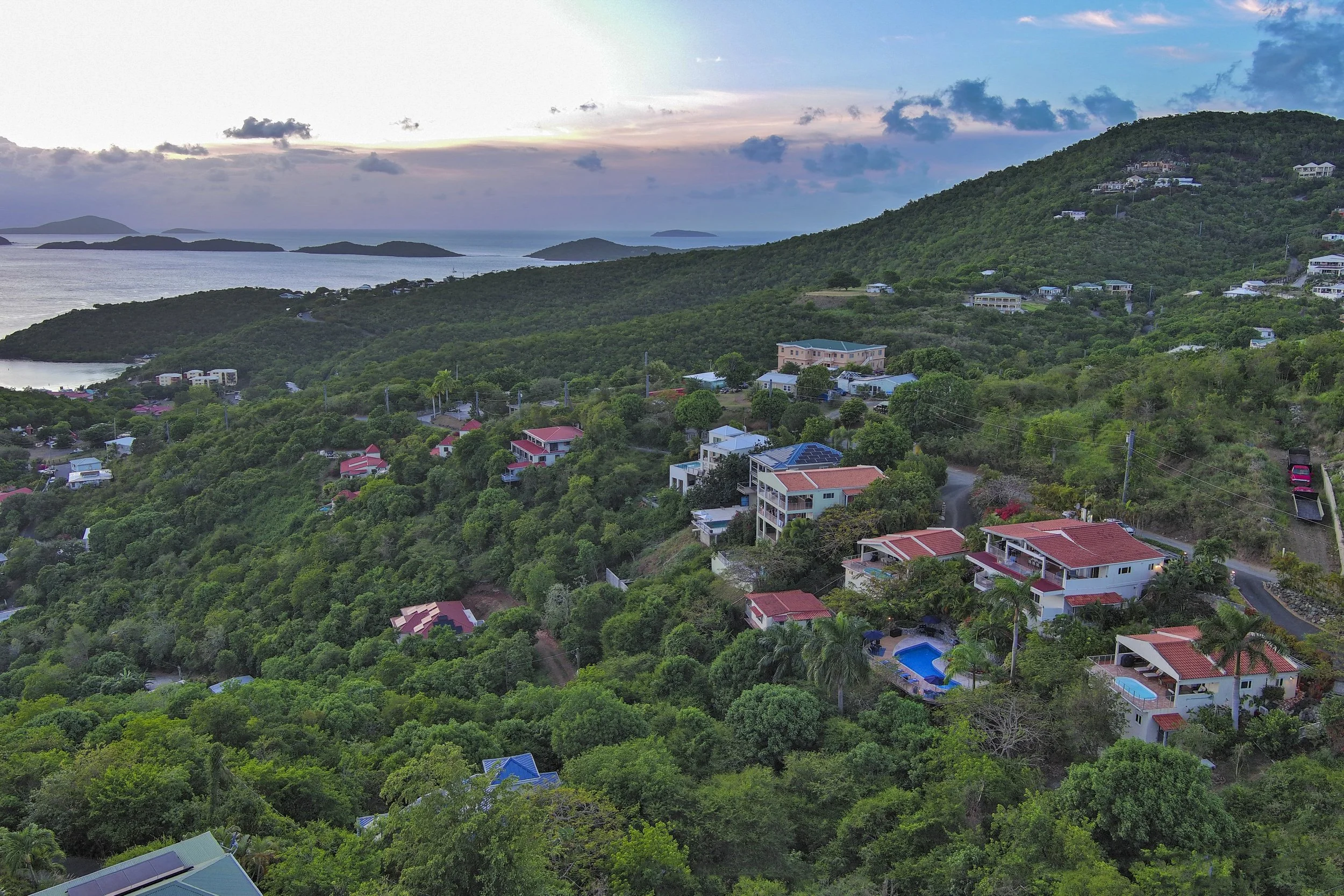 Mandavilla St. John USVI vacation rental Landscape view of a coastal hillside with residential houses, lush green trees, and distant islands in the ocean under a partly cloudy sky at sunset.