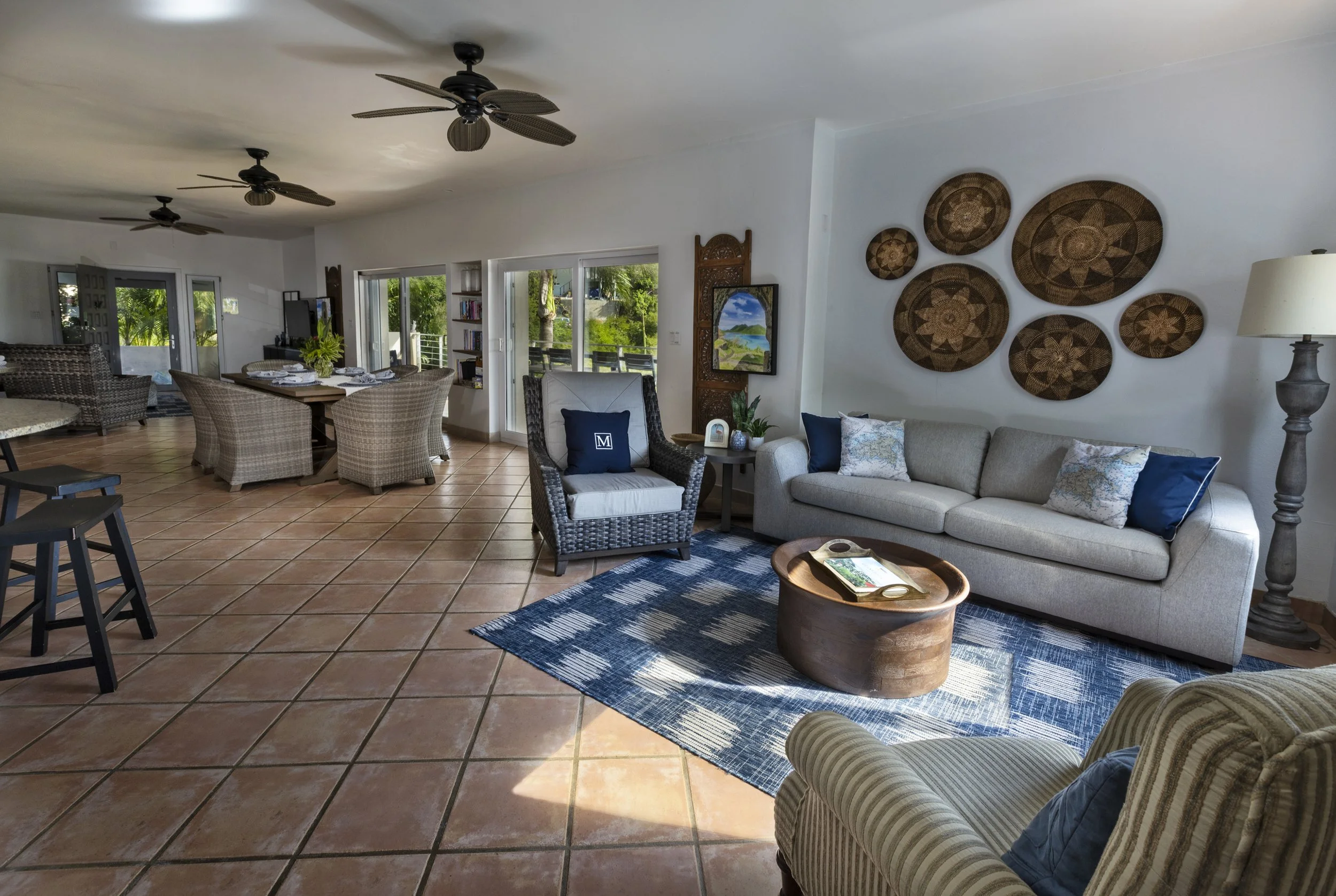 Open-plan living and dining area with wicker chairs, a beige sofa with blue pillows, a wooden coffee table, and decorative wall art made of woven baskets. Large sliding glass doors lead to a balcony with greenery outside. Ceiling fans are on the ceil