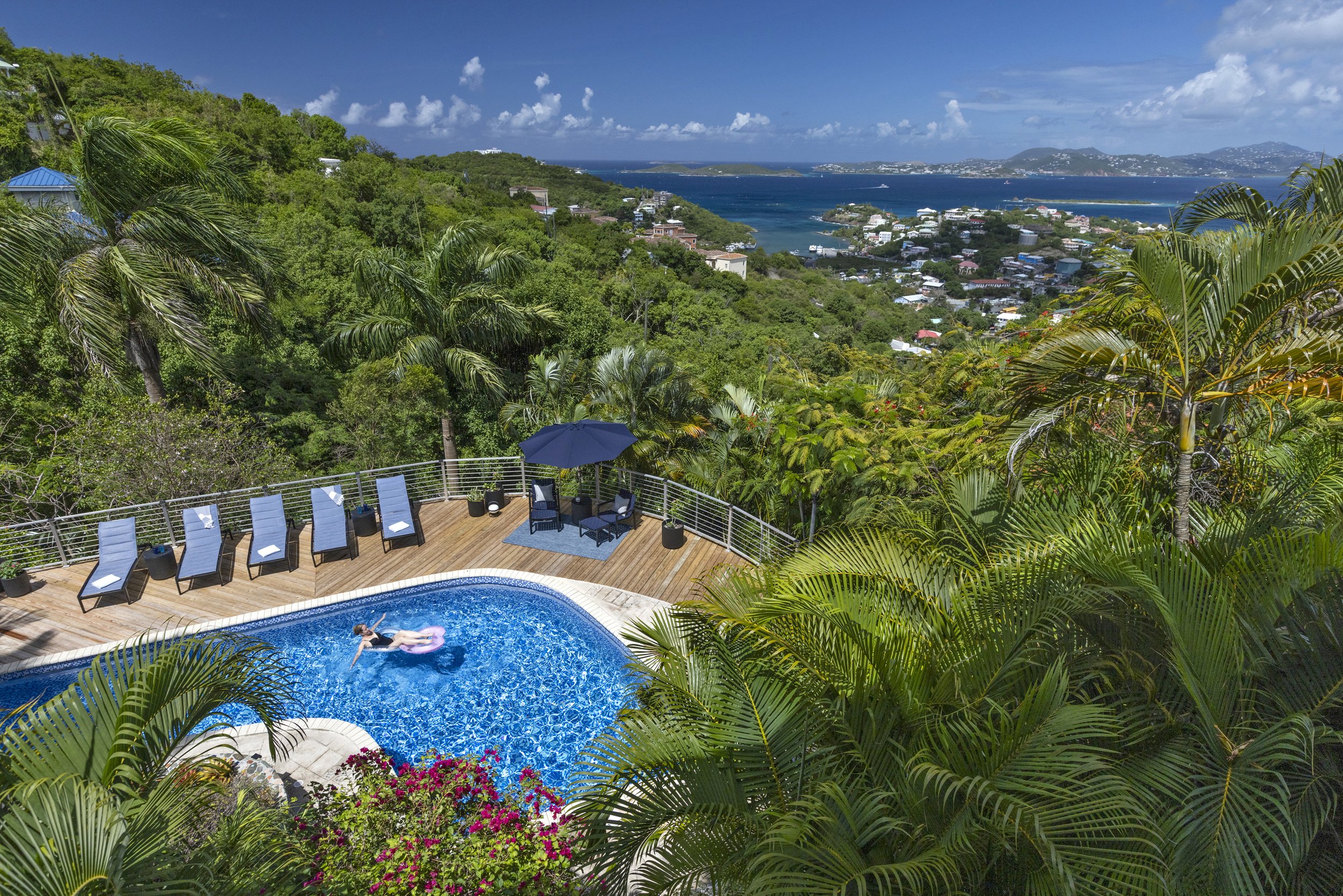 A view of a hillside with lush green vegetation overlooking a bay with boats and buildings in the distance. In the foreground, an outdoor swimming pool with a person floating on an inflatable, surrounded by lounge chairs and umbrellas.