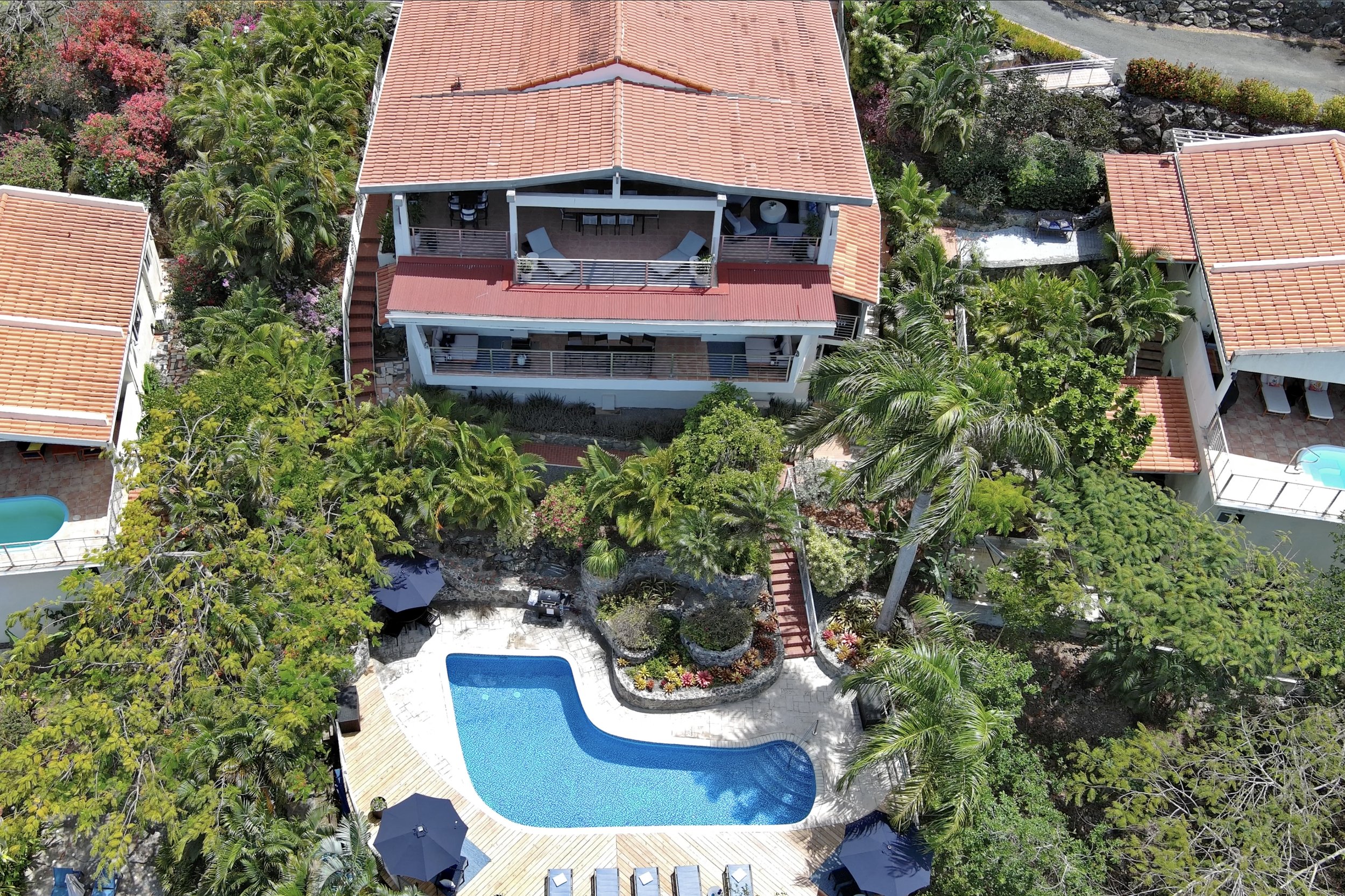 Mandavilla St. John USVI vacation rental Aerial view of a house with a red-tile roof, surrounded by lush green trees and plants. There are outdoor seating areas, a swimming pool, and a deck with umbrellas.