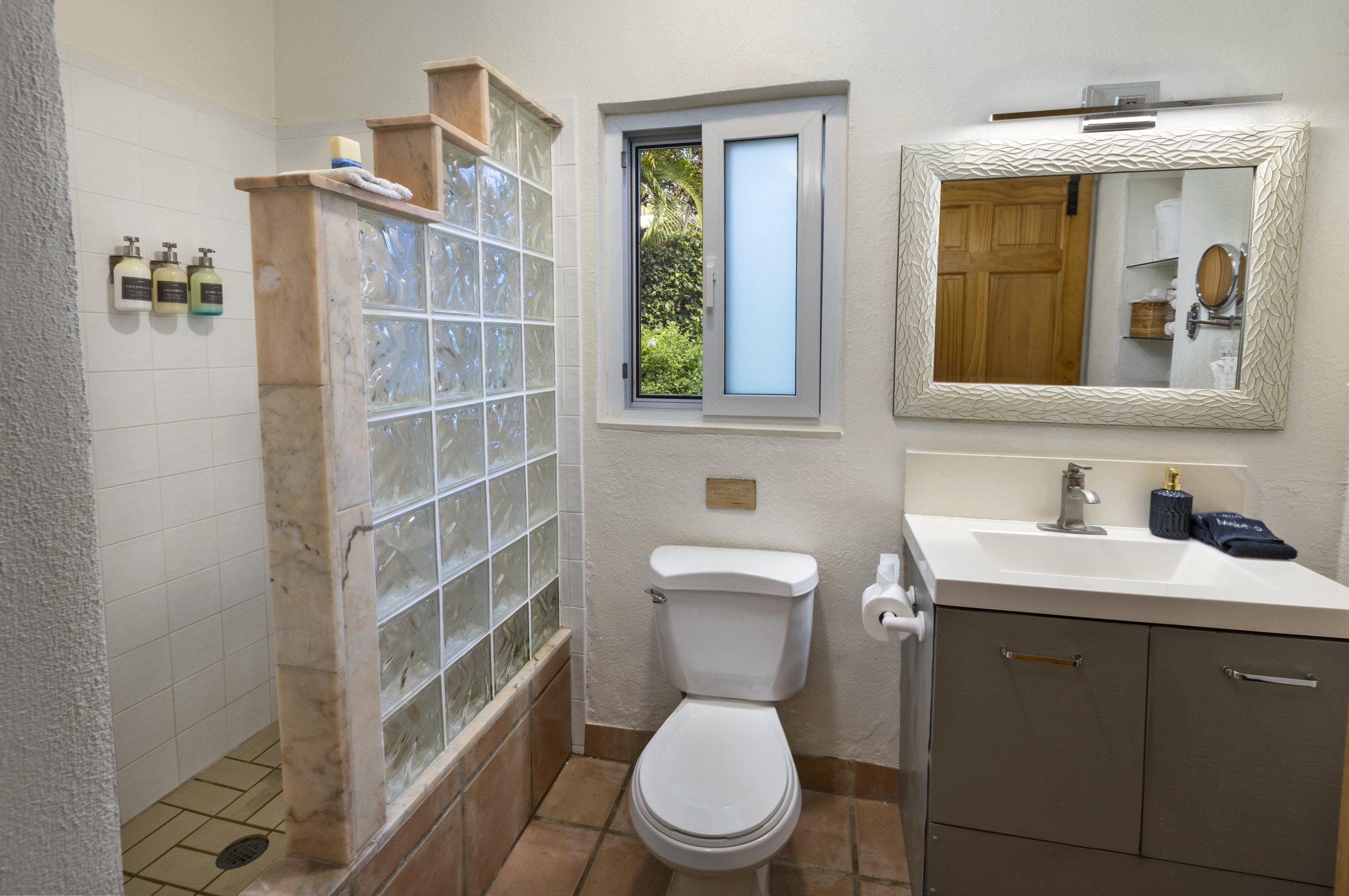 A small bathroom with a glass block wall dividing the shower area from the toilet and vanity. There is a window with a view of greenery, a rectangular mirror with a textured frame, a white sink with a dark cabinet beneath, and shelves with towels and