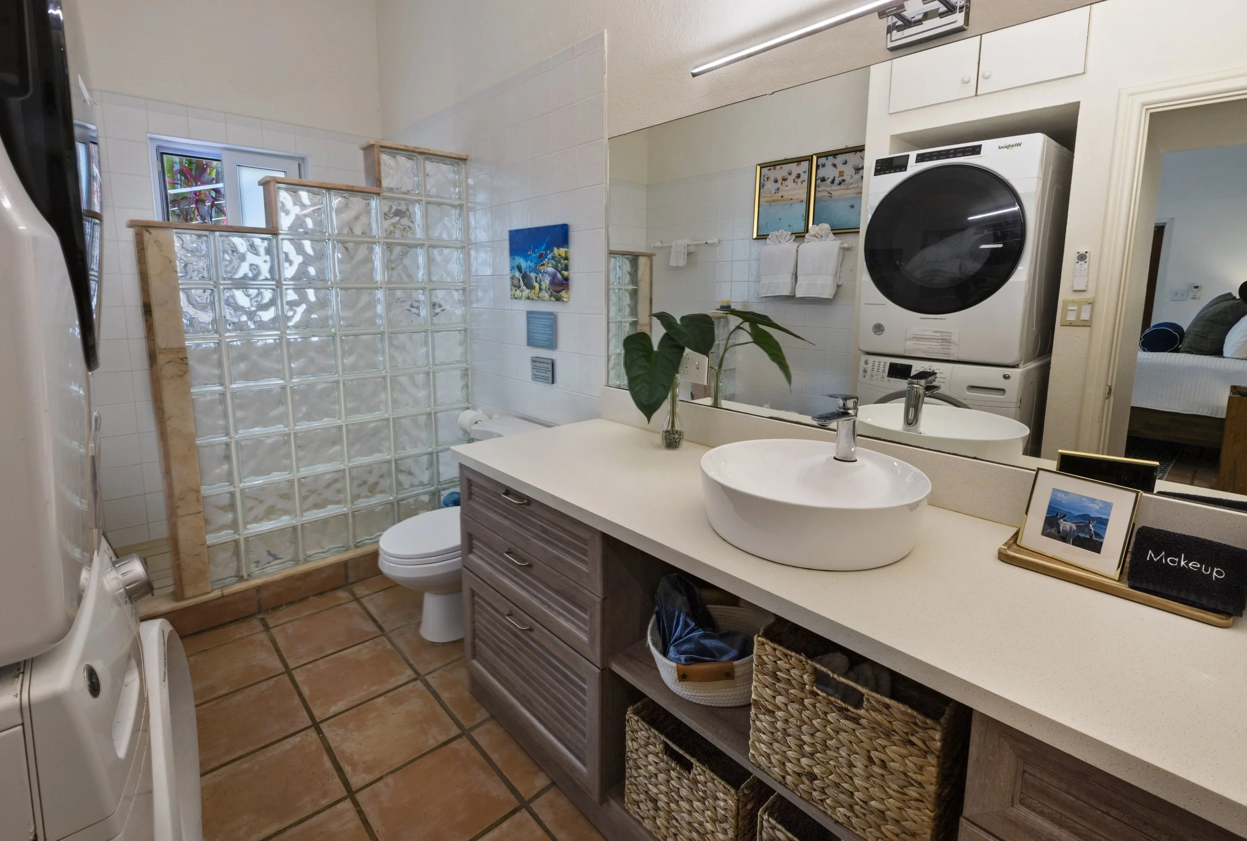A bathroom with a glass block shower, white tiled walls, a white sink with a large mirror, and a stacked washer and dryer. Decorations include framed beach pictures, a plant, towels, a small framed photo, and baskets for storage. The bathroom has a s