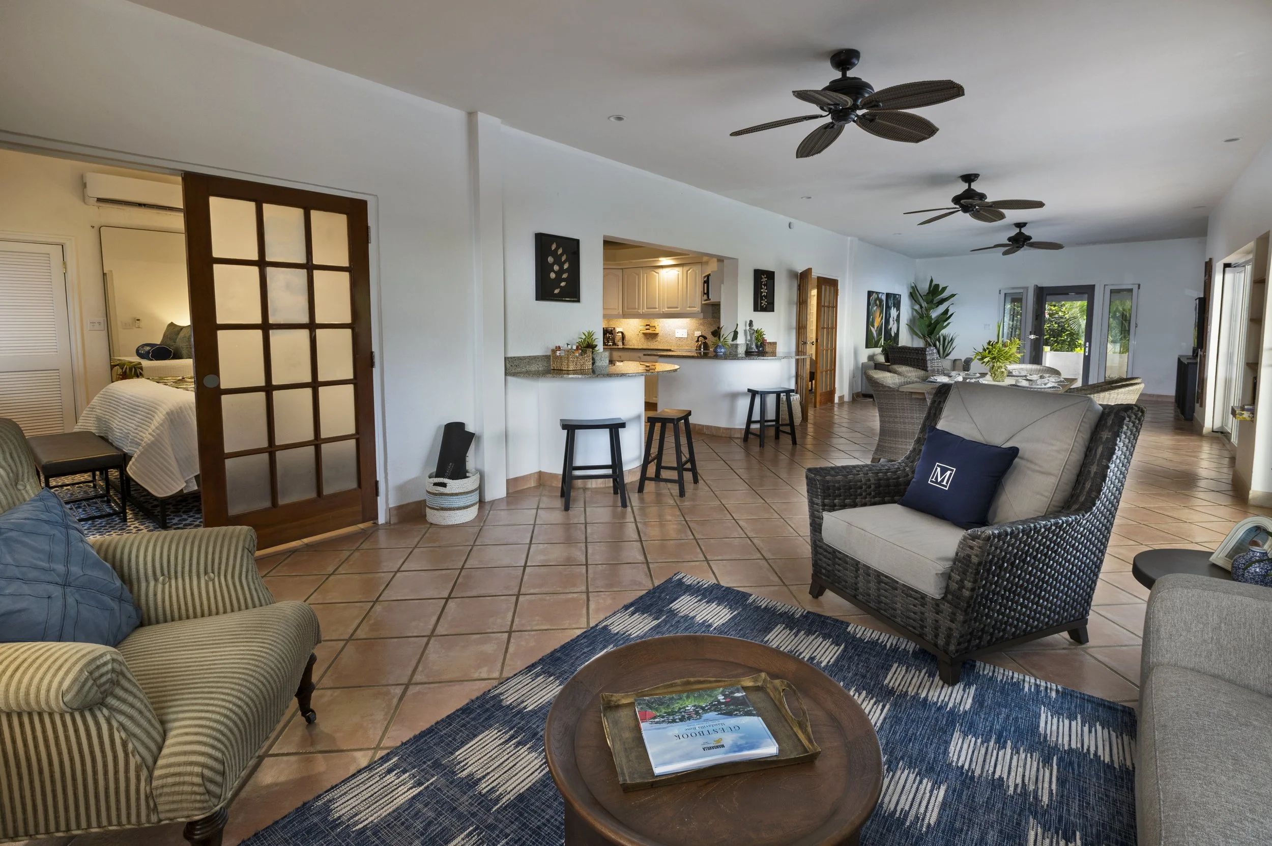 Living room with tiled floor, wicker and upholstered chairs, a round wooden coffee table with a magazine, and a dark blue rug. There is a dining area with a table and chairs, and a kitchen with a breakfast bar and stools. Ceiling fans and potted plan
