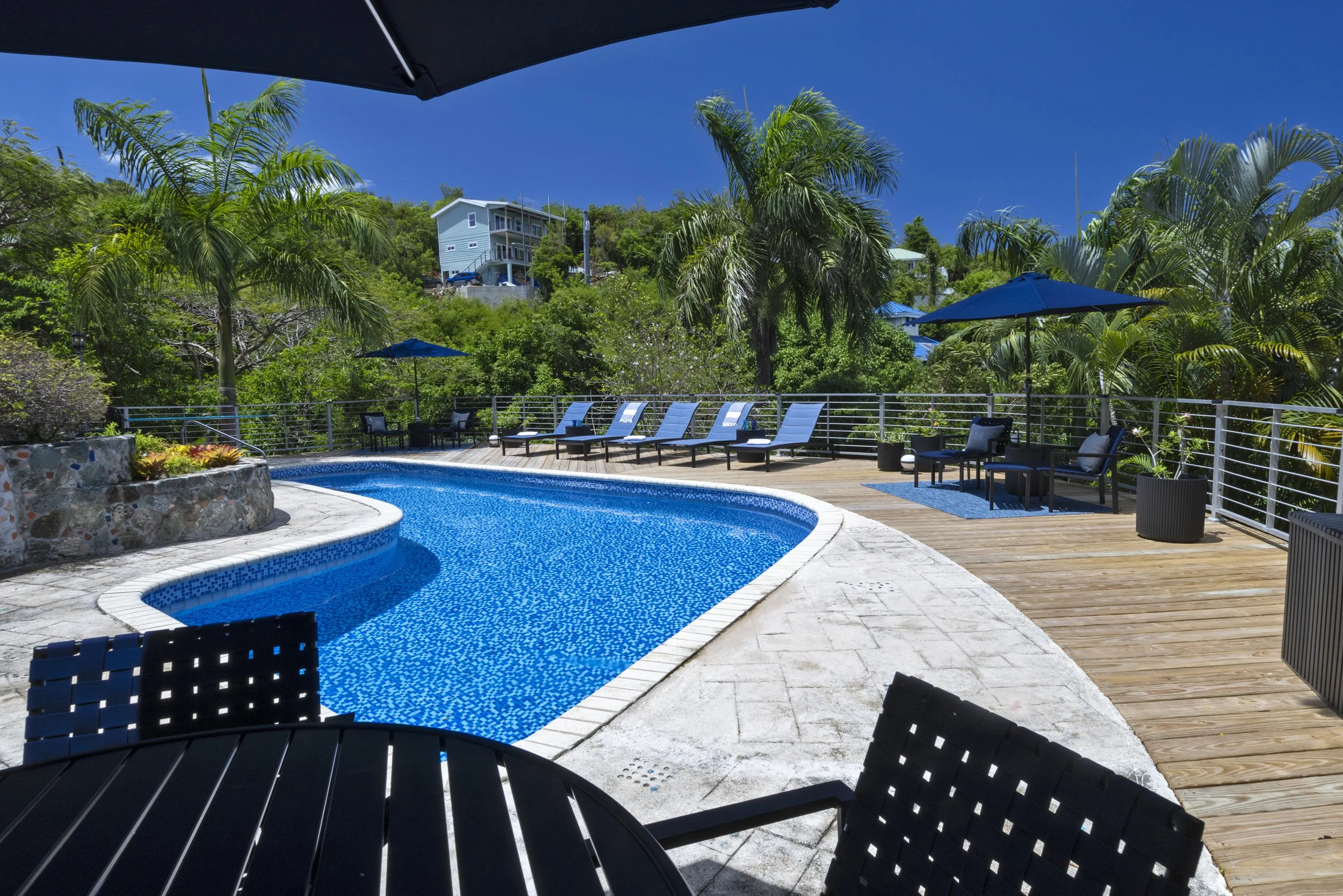 Mandavilla St. John USVI vacation rental pool area with a blue mosaic-tiled pool, surrounded by a wooden deck, lounge chairs, umbrellas, and lush green palm trees, with a hillside clear blue sky ocean view.