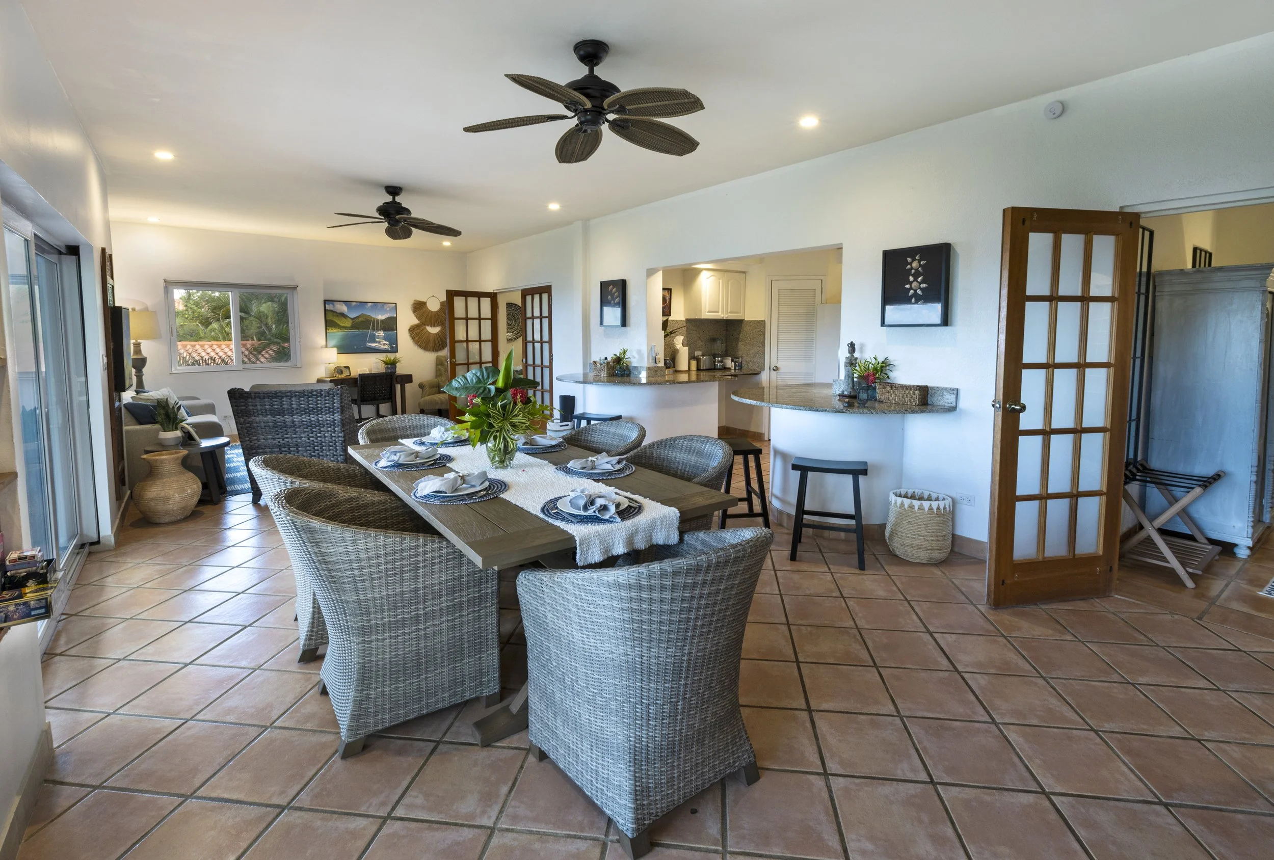 A spacious, open-concept living and dining area with tiled flooring, wicker chairs around a wooden dining table with a floral centerpiece, a living area with a sofa, lamps, and wall art, and a view into a kitchen with white cabinets and granite count