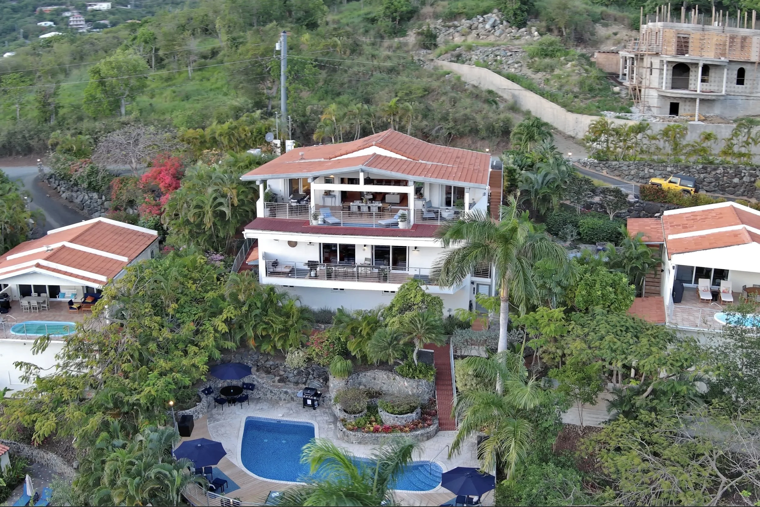 Mandavilla St. John USVI vacation rental Aerial view of a hillside residential area with houses, greenery, a swimming pool with umbrellas, and ongoing construction on a multi-story building in the background.