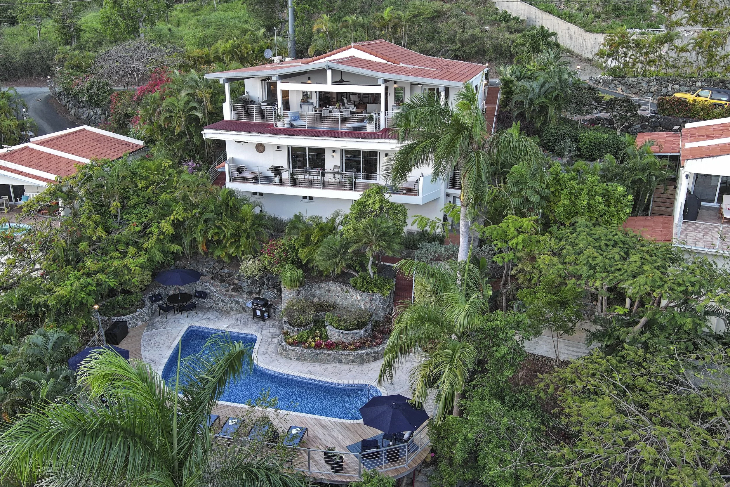 Mandavilla St. John USVI vacatio multi-story house with red tiled roofs surrounded by lush greenery, including palm trees and flowering bushes. There is a backyard with a swimming pool, patio furniture, umbrellas, and a fire pit area.