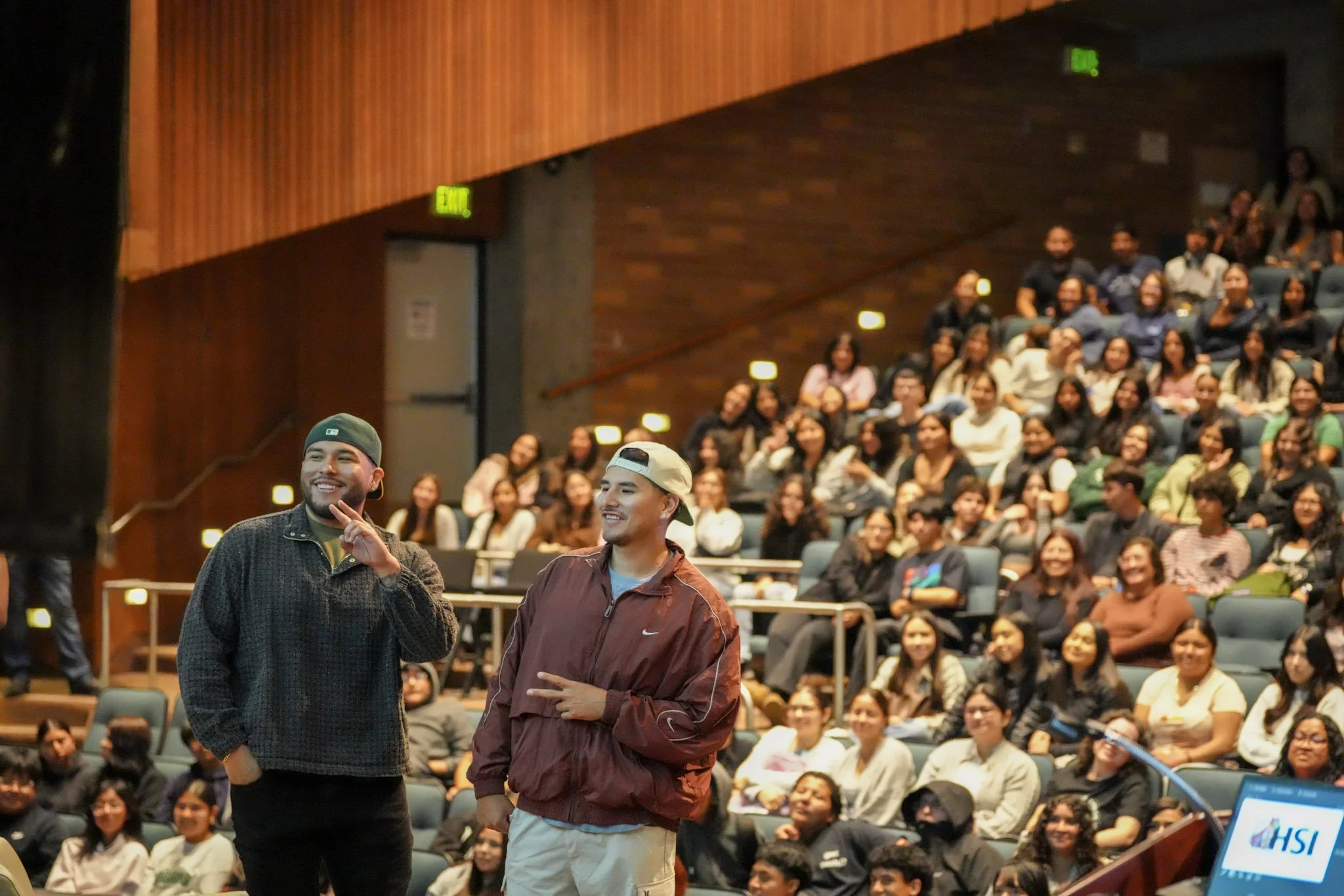 Two young men standing in front of an audience in a conference room with wooden walls and green exit signs. The audience appears engaged and smiling.