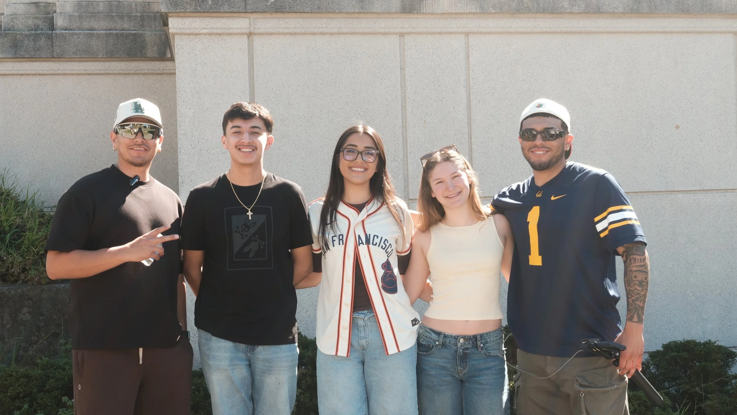 Group of five young adults standing outside against a plain wall, smiling at the camera, dressed casually, with some wearing sunglasses and hats.