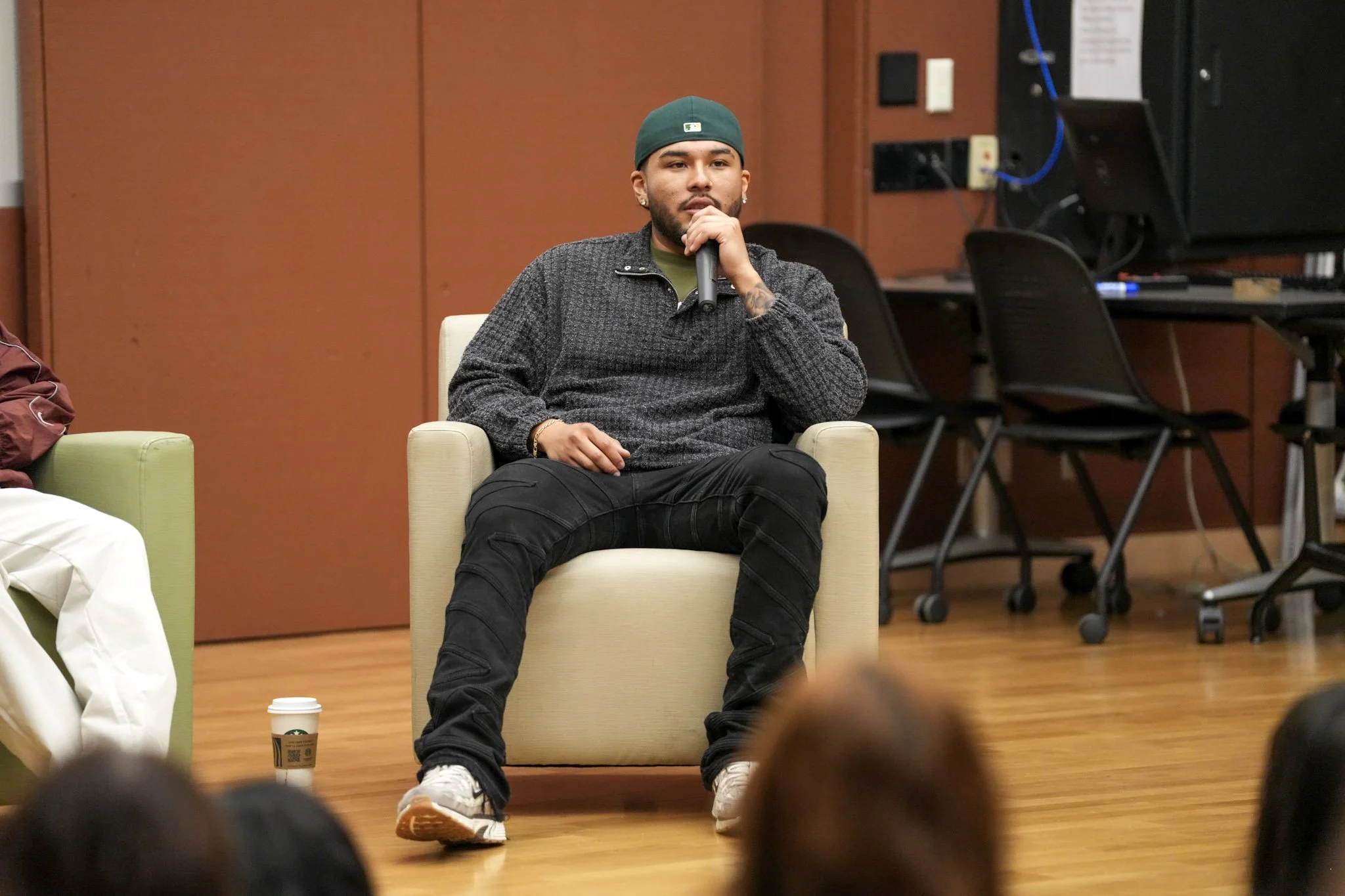 A young man with a beard, earrings, and a green baseball cap is sitting in a cream armchair, speaking into a microphone, in a room with chairs, tables, and a large monitor.