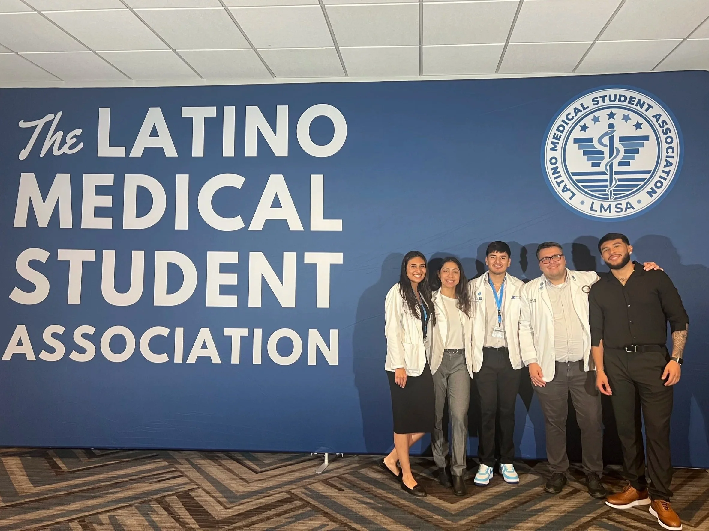 Group of five people posing in front of a blue wall with a logo for the Latin Medical Student Association. The group includes three men and two women, all dressed in white or black professional attire, smiling at the camera.