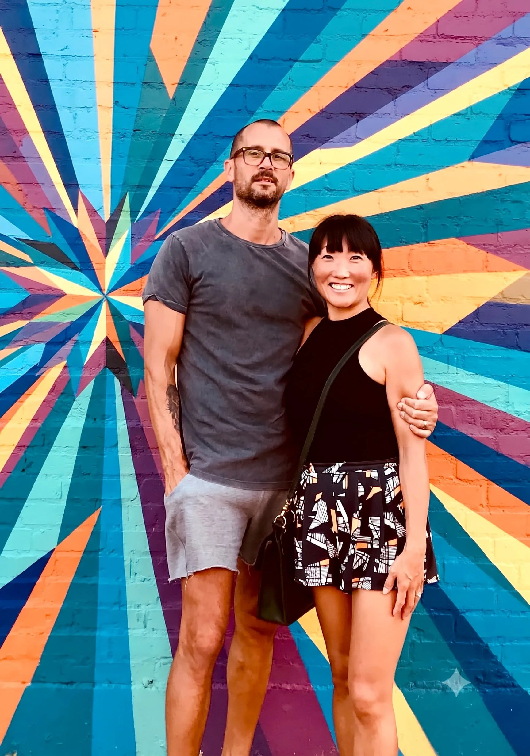 Bentonville, AR personal trainer Mark Saint, owner of Mark Saint Personal Training, stands with his wife in front of a vibrant, geometric brick wall mural.