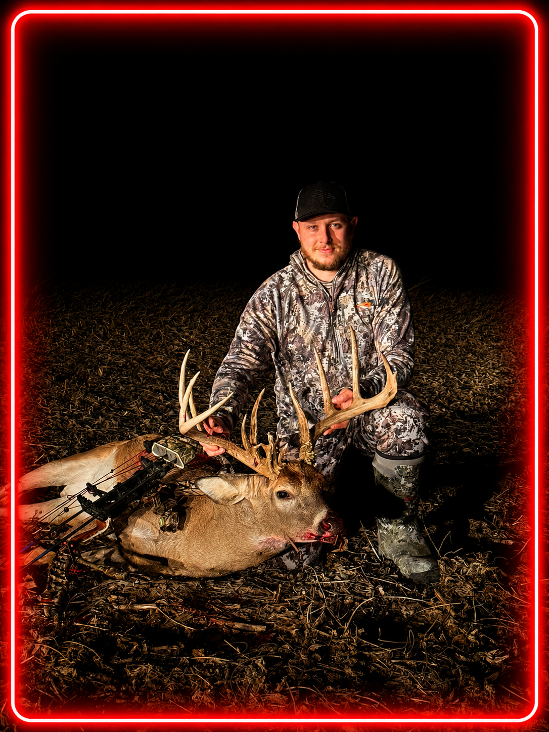 A man in camouflage outfit kneeling on soil at night, holding a hunted deer with large antlers, with a bow and arrow nearby, illuminated by red neon border.