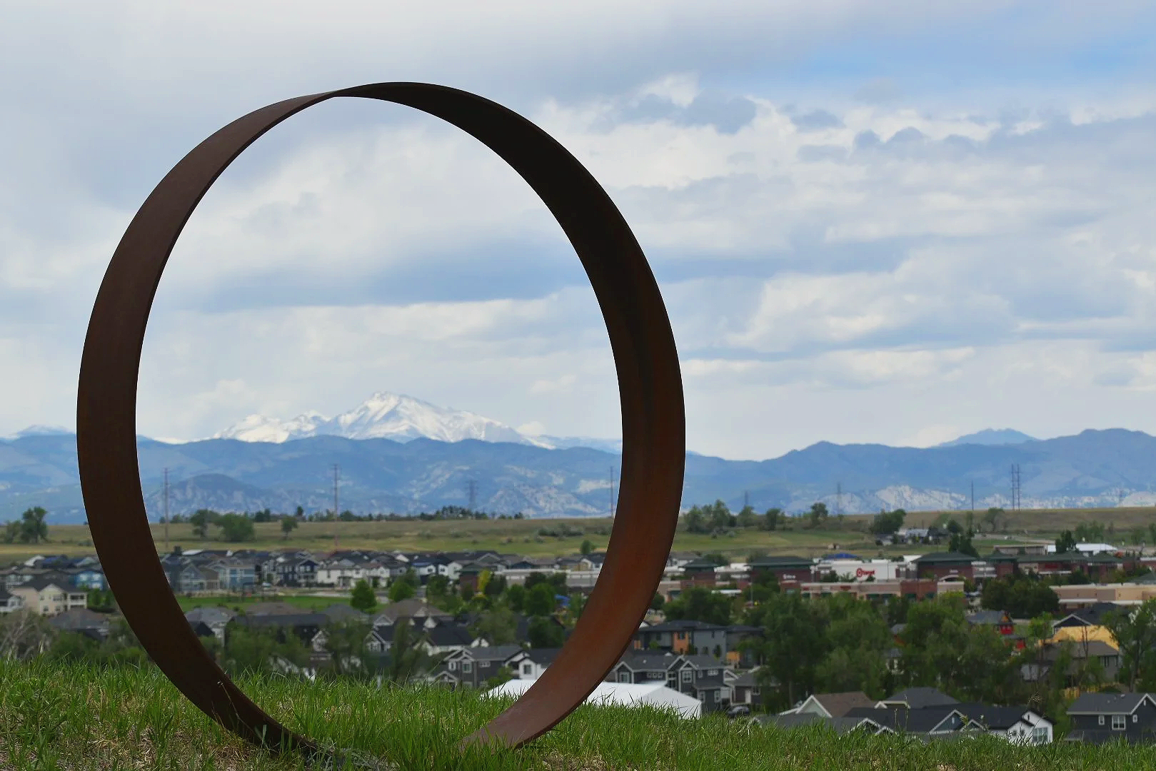 A large, rust-colored metal sculpture shaped like a circle with an opening in the center, situated on a grassy hill. Behind it, there are mountains with snow-capped peaks and a town with houses and buildings.