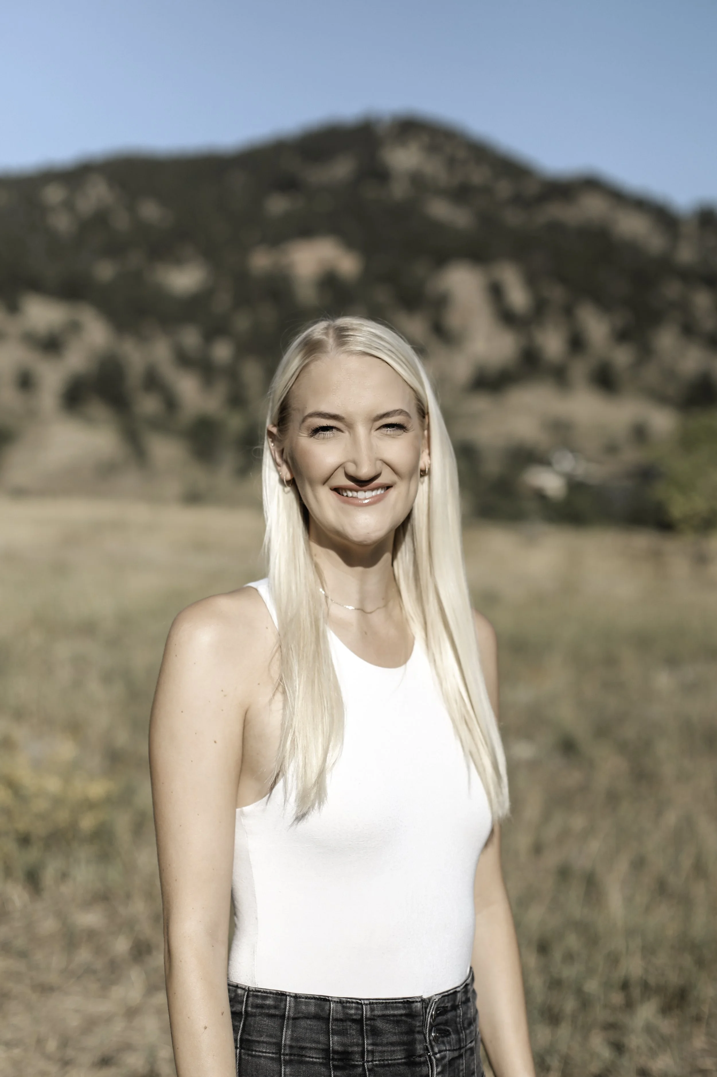 A young blonde woman smiling outdoors with a mountain in the background, wearing a white tank top and black jeans.