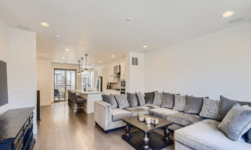 Open-concept living room and kitchen in a modern apartment with gray sectional sofa, throw pillows, coffee table, kitchen island, hanging pendant lights, stainless steel appliances, and balcony with outdoor furniture.