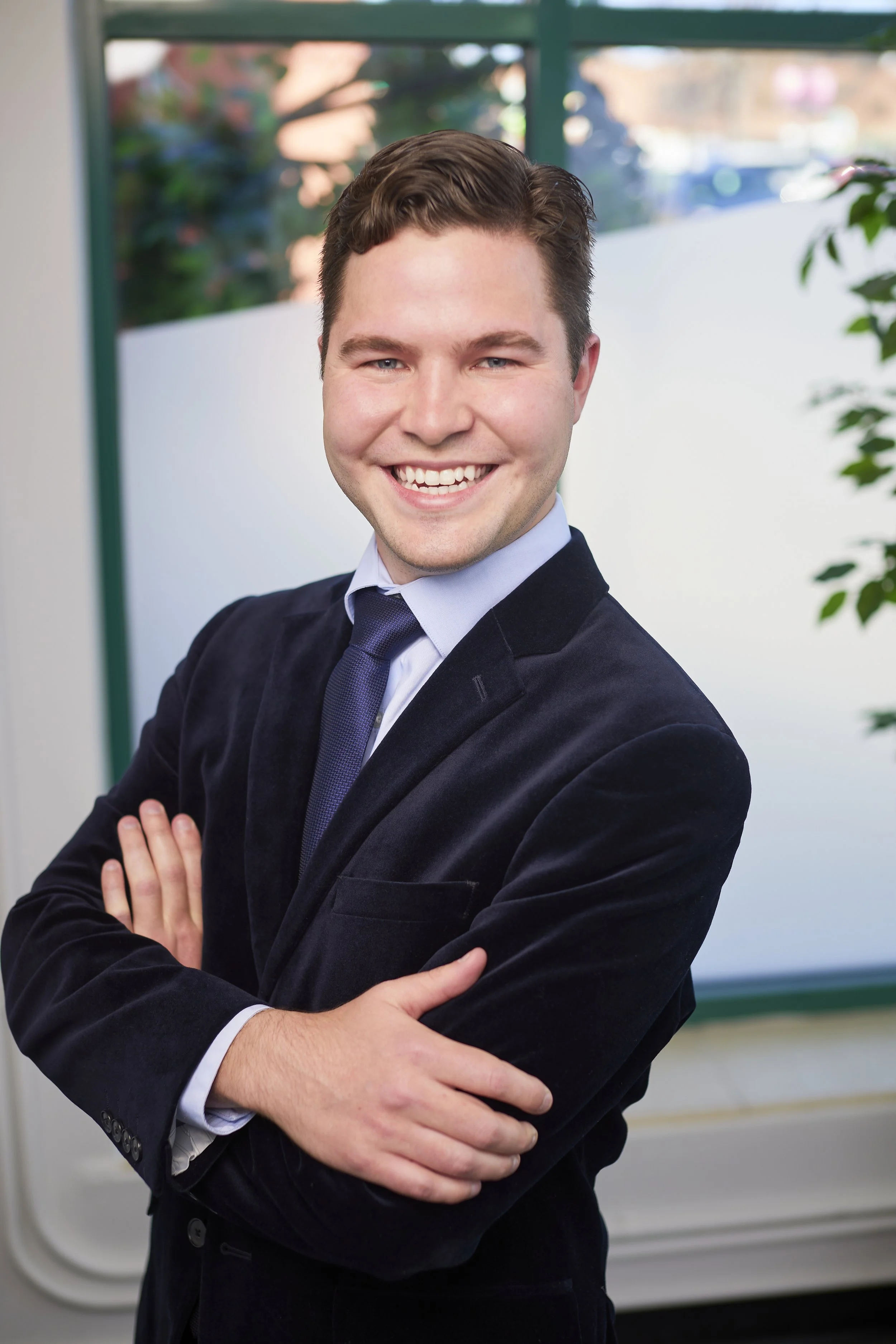 A young man in a suit smiling with arms crossed in a professional setting, with large windows and plants in the background.